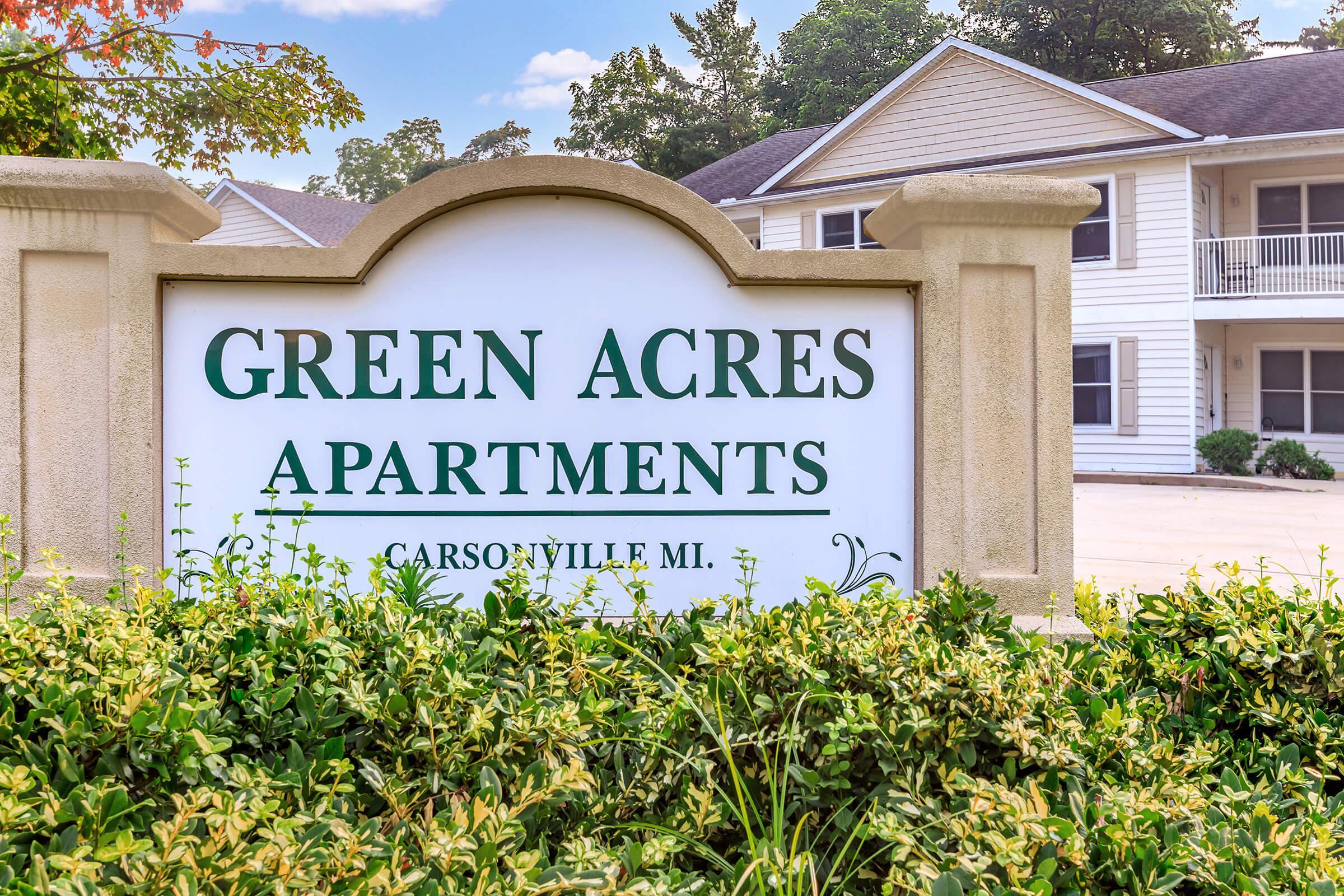 Sign for Green Acres Apartments located in Carsonville, MI, surrounded by green bushes and trees. The sign features a clean, modern design with bold lettering that emphasizes the name of the apartments. In the background, there is a glimpse of the apartment buildings.