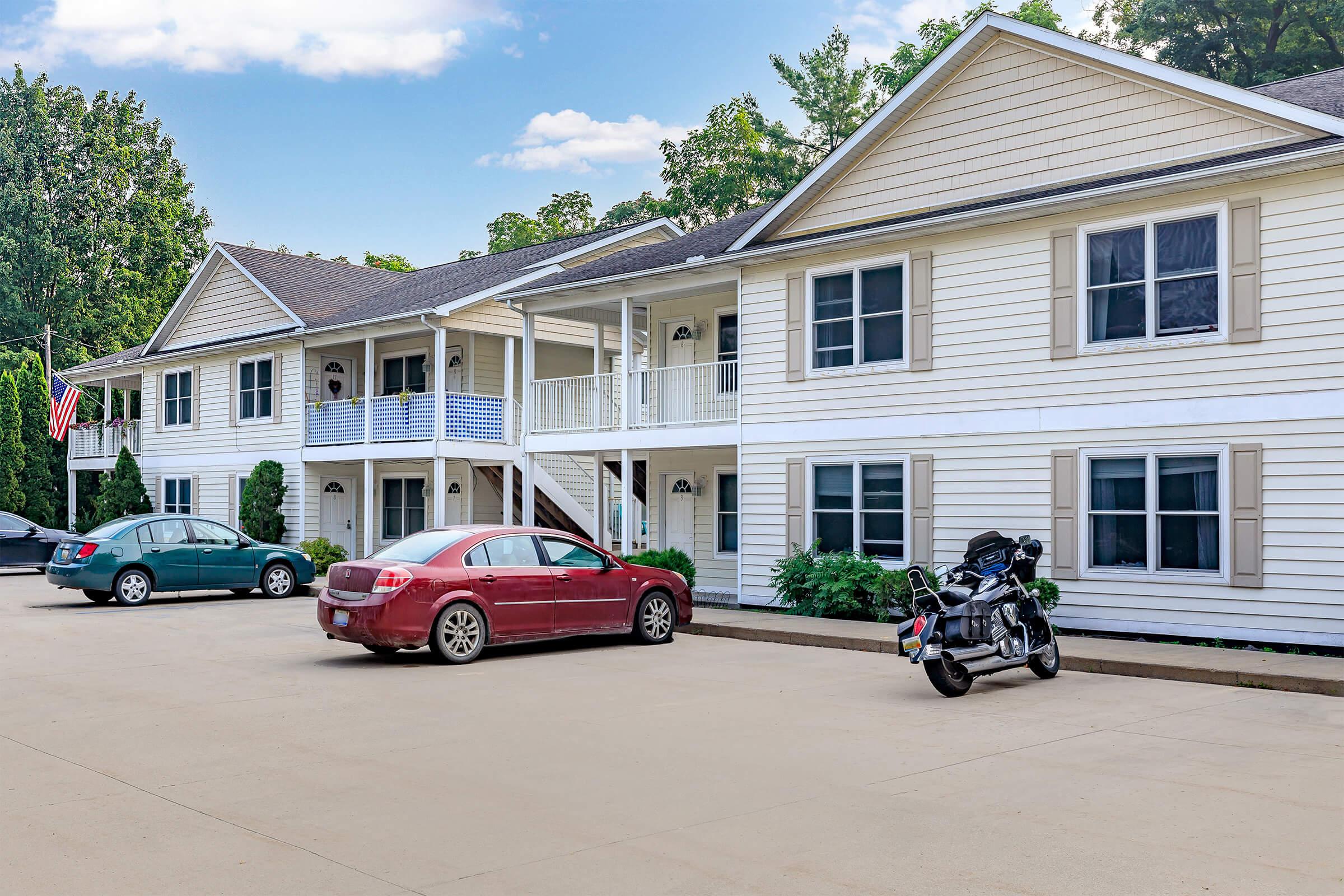 A two-story building with a white exterior and blue accents, featuring apartment-style entrances. In the foreground, there are two parked cars: a silver sedan and a red sedan, along with a motorcycle. The area is landscaped with greenery, and an American flag is visible in the background.