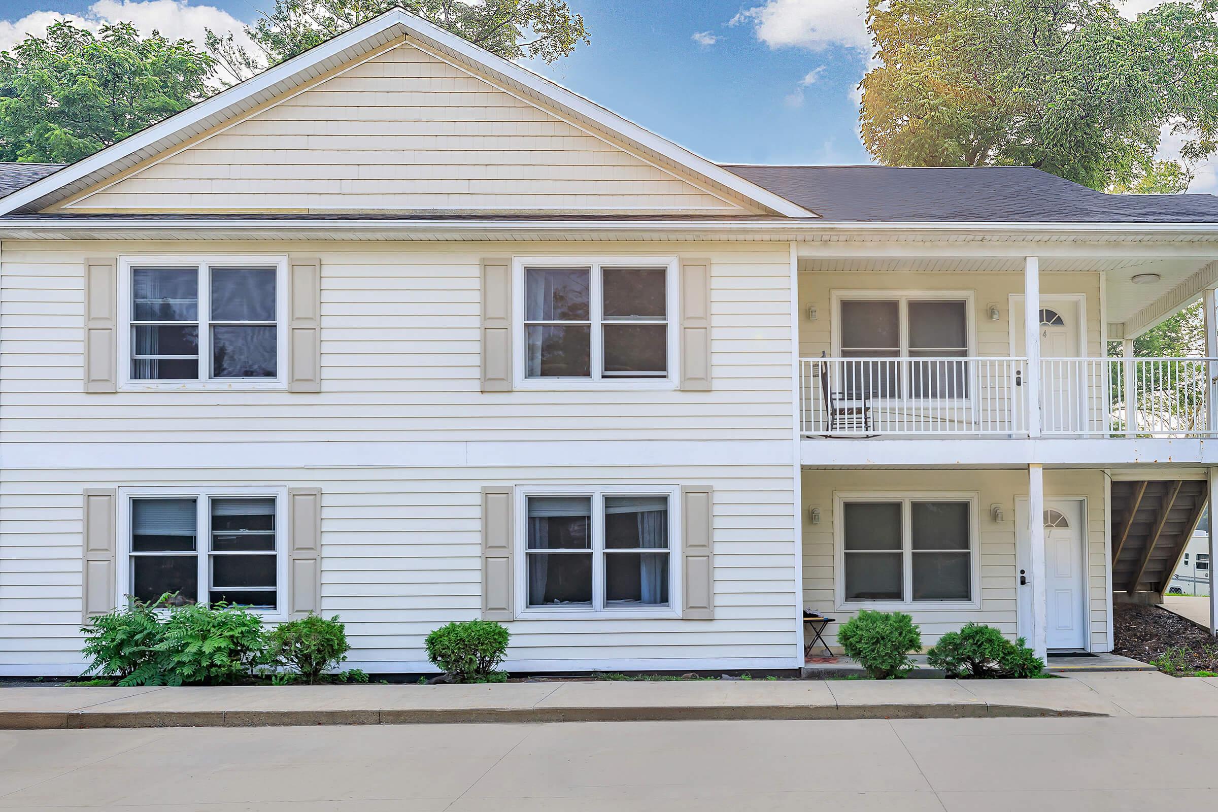 Two-story residential building with white siding and light beige shutters. The building features a small balcony on the upper floor, surrounded by greenery. Windows are evenly spaced, and there is a front door on the right side. The setting appears friendly and well-maintained, under a bright sky with some clouds.