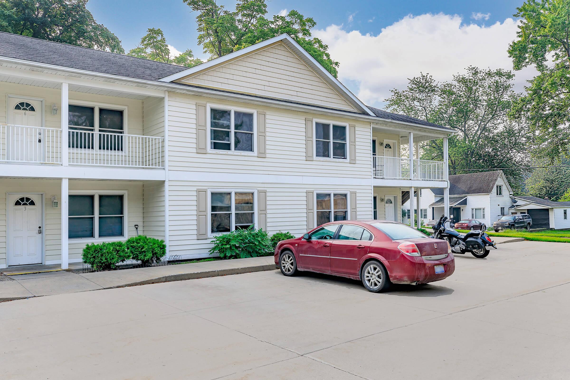 A two-story white building with a porch and multiple windows, set in a residential area. In the foreground, a red sedan is parked next to a motorcycle. Lush greenery is around the building, and other houses are visible in the background under a partly cloudy sky.