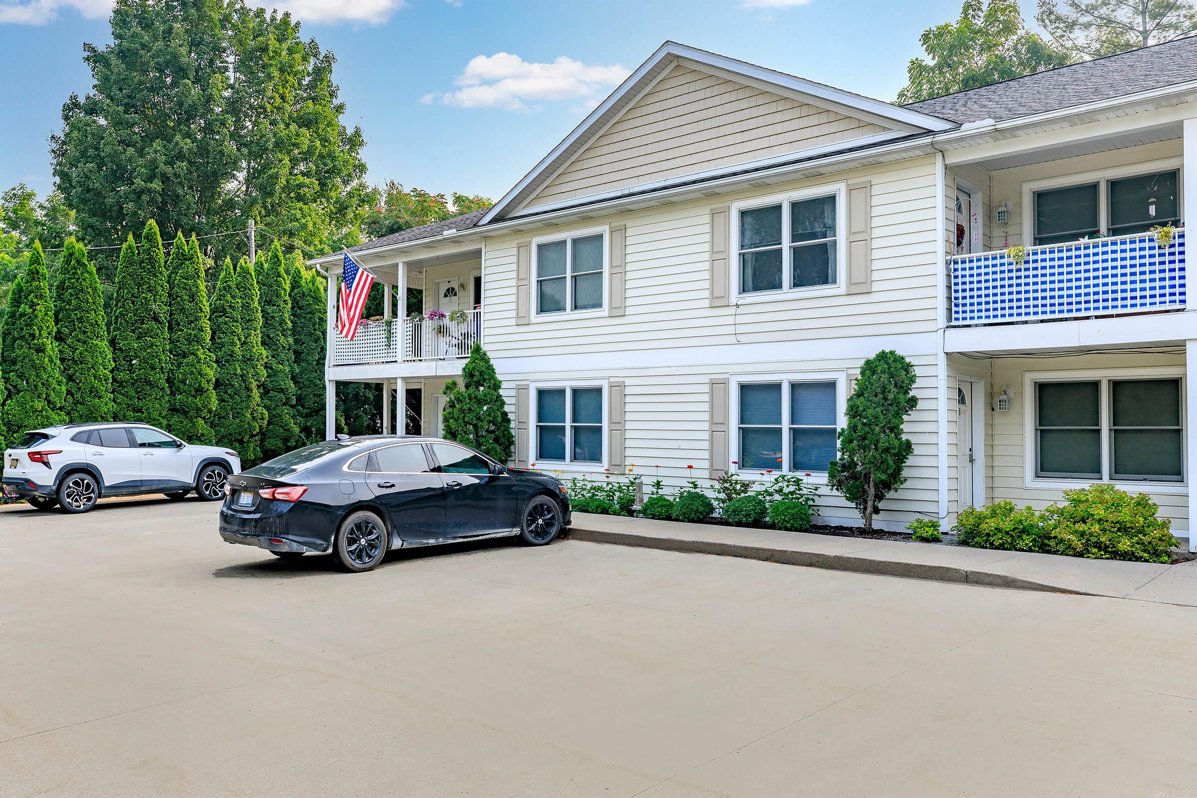 Exterior view of a two-story residential building with white siding and a U.S. flag hanging from the balcony. There are neatly trimmed trees and shrubs in the front yard. A black car is parked in the driveway, alongside another vehicle, with a clear sky in the background.