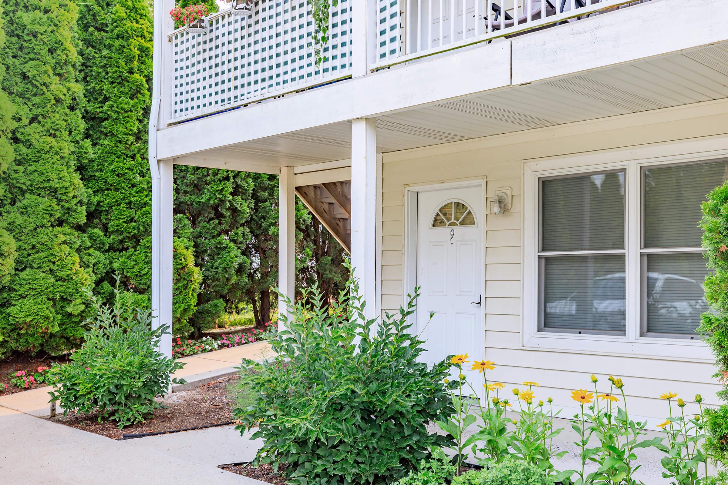 A peaceful exterior view of a home featuring a white door, green bushes, and blooming yellow flowers. The house is two stories with a balcony above and well-maintained landscaping. Lush green trees frame the scene, providing a serene and inviting atmosphere.