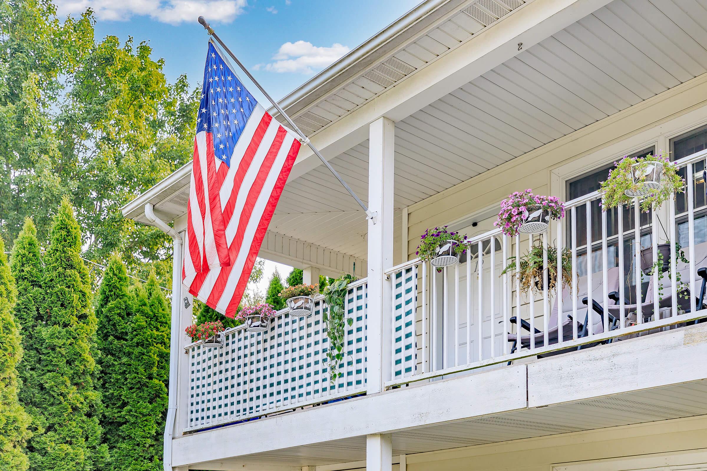 A large American flag hanging from a porch railing, surrounded by lush green trees. The porch features white railings with several flower pots and chairs, creating a welcoming outdoor space. The house has a light-colored exterior, and the sky is bright with a few clouds.