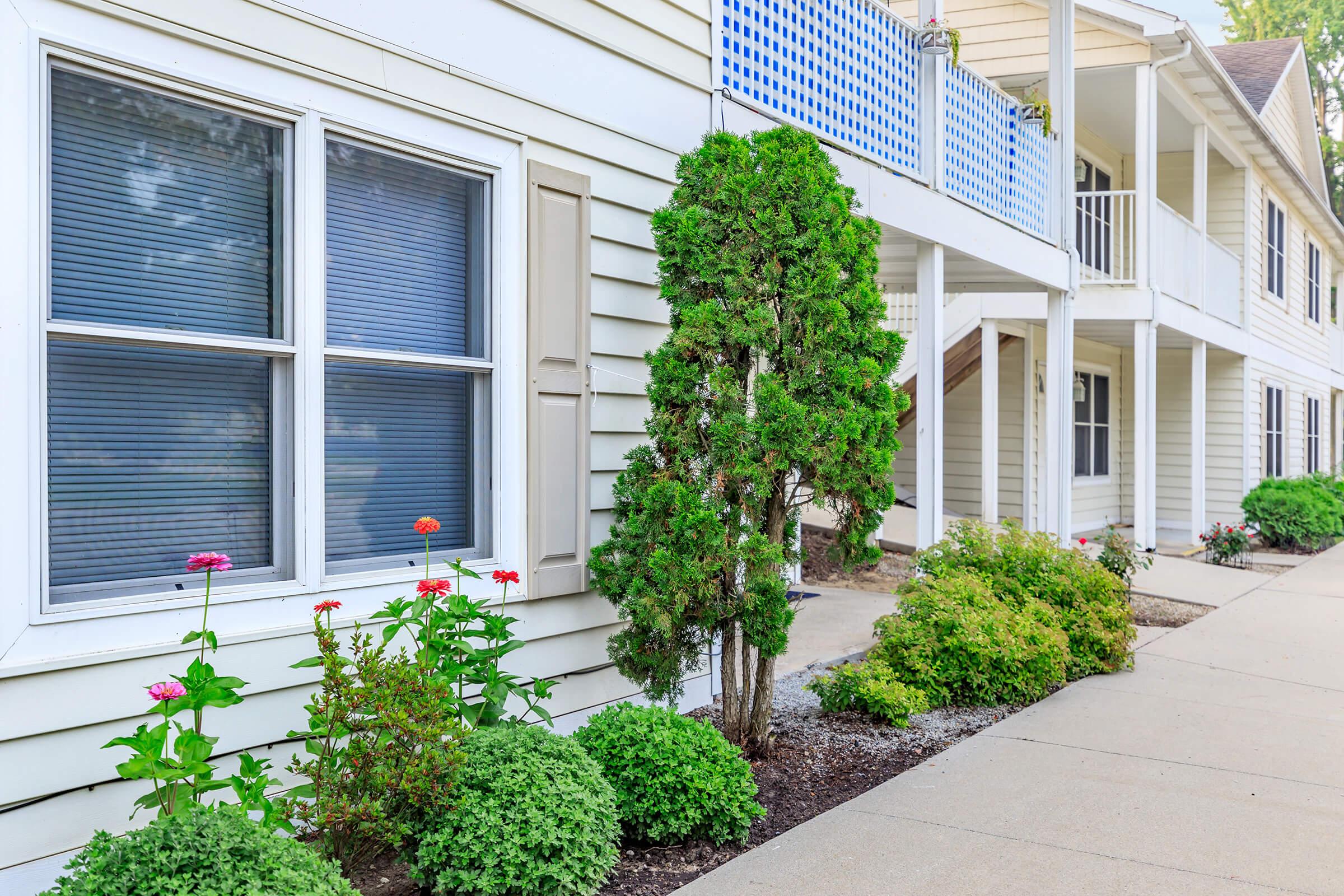 A well-maintained exterior of a residential building featuring white siding. In the foreground, colorful flowers and neatly trimmed shrubs line the walkway, with two windows visible on the left side. The entrance area has steps leading to a porch, adding to the inviting atmosphere.