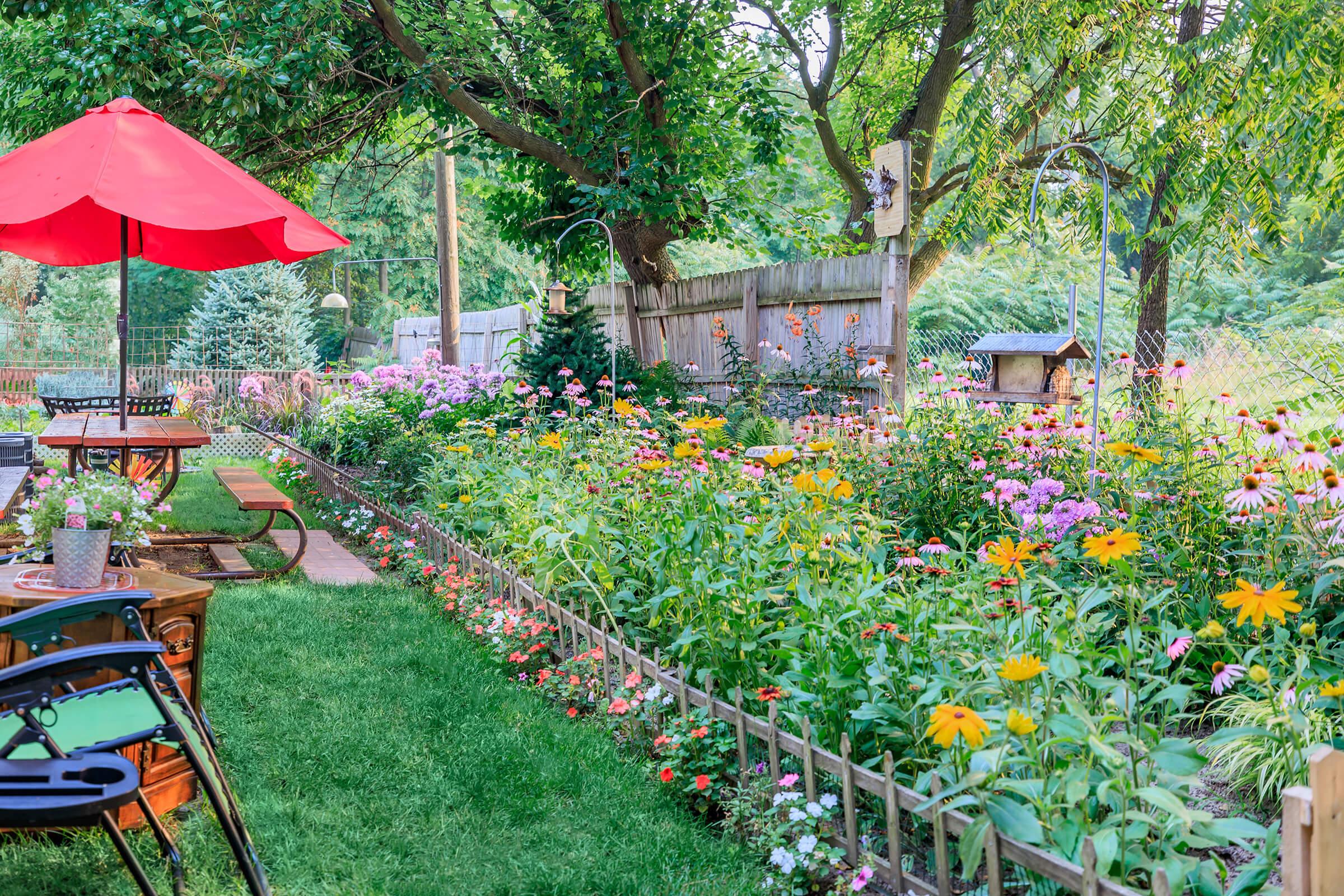 A vibrant garden filled with colorful flowers, including coneflowers and daisies, alongside a manicured lawn. A patio area features wooden furniture and a red umbrella, while a wooden fence and trees create a serene backdrop. Bird feeders and garden decorations add charm to the scene.