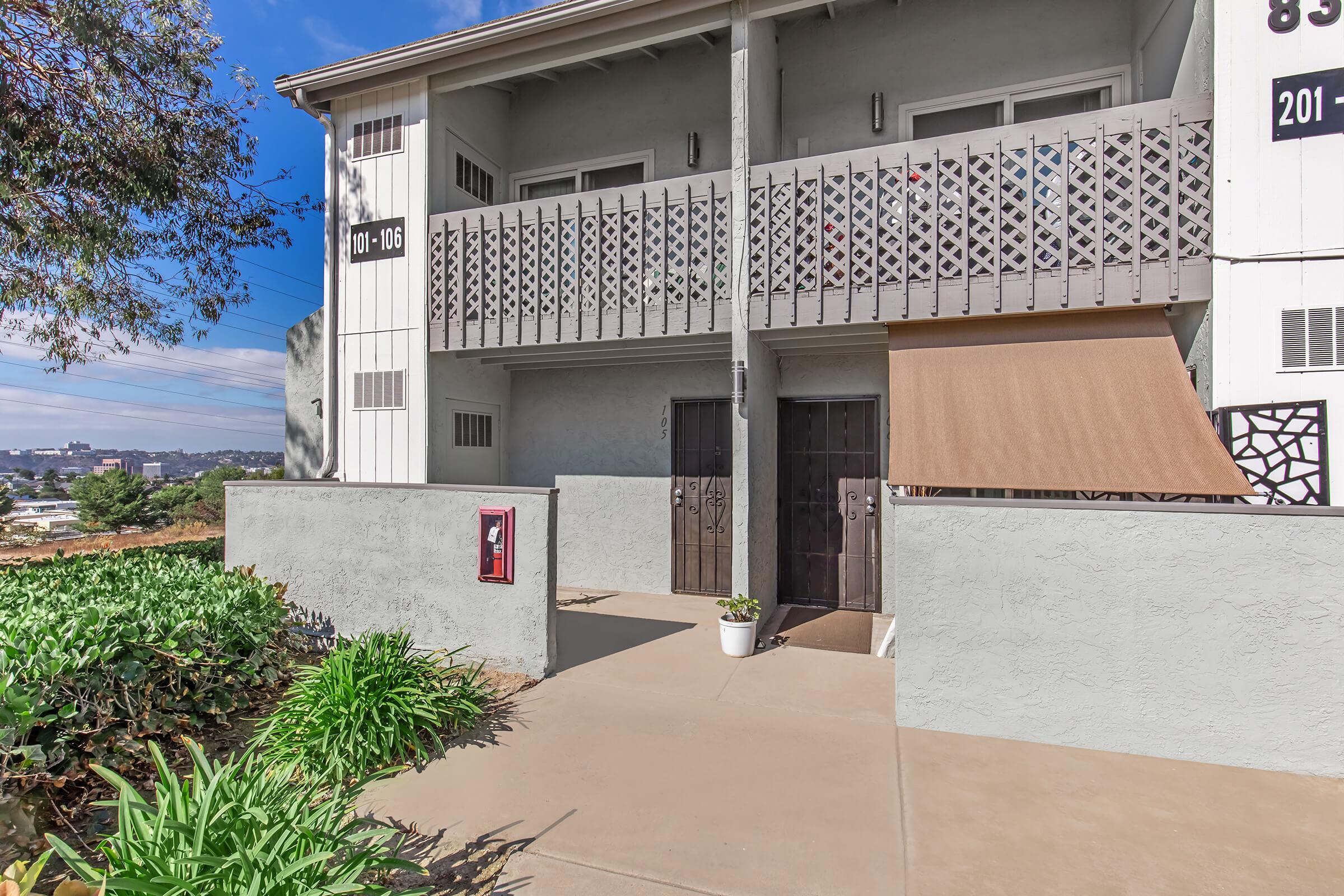 Exterior view of an apartment building featuring a balcony with a decorative railing, two entry doors, and a small potted plant beside a concrete pathway. The surrounding area includes greenery and a clear blue sky in the background.