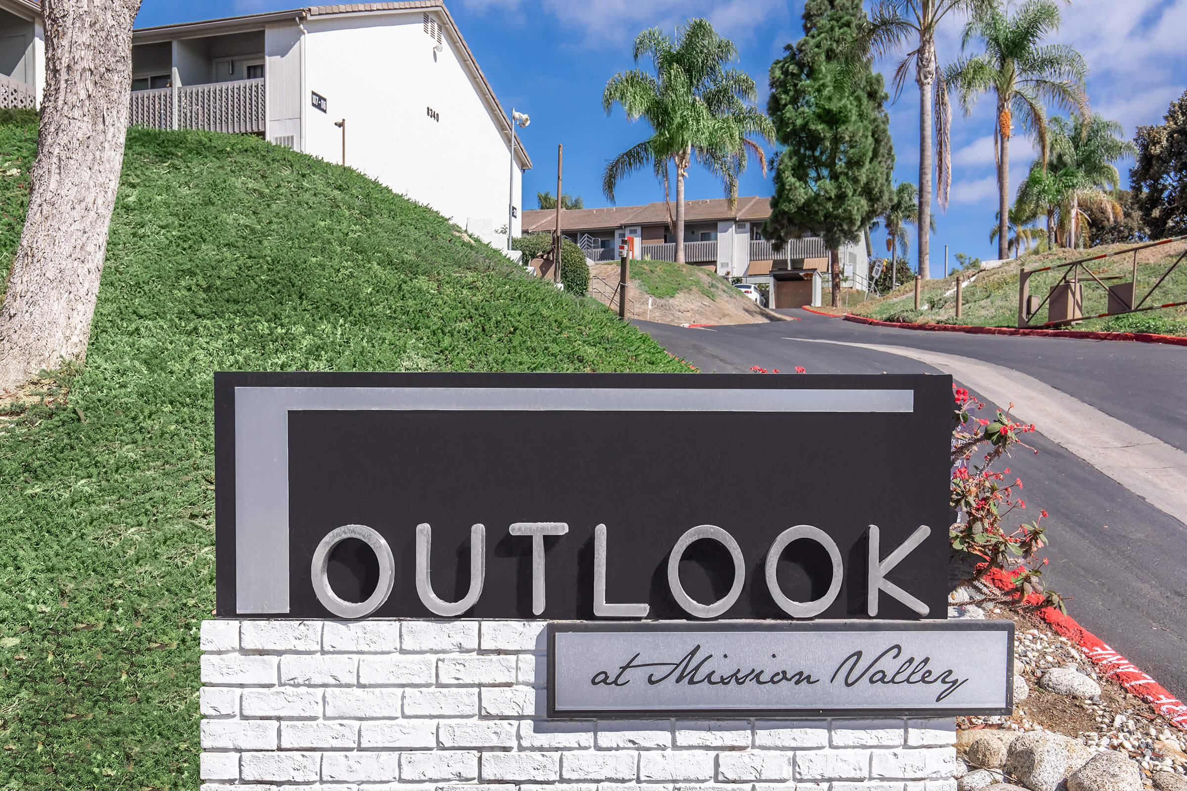 Sign for "OUTLOOK at Miron Valley," featuring modern typography on a landscaped area with trees and a residential complex in the background. The scene has a clear blue sky and a paved road leading up to the buildings.