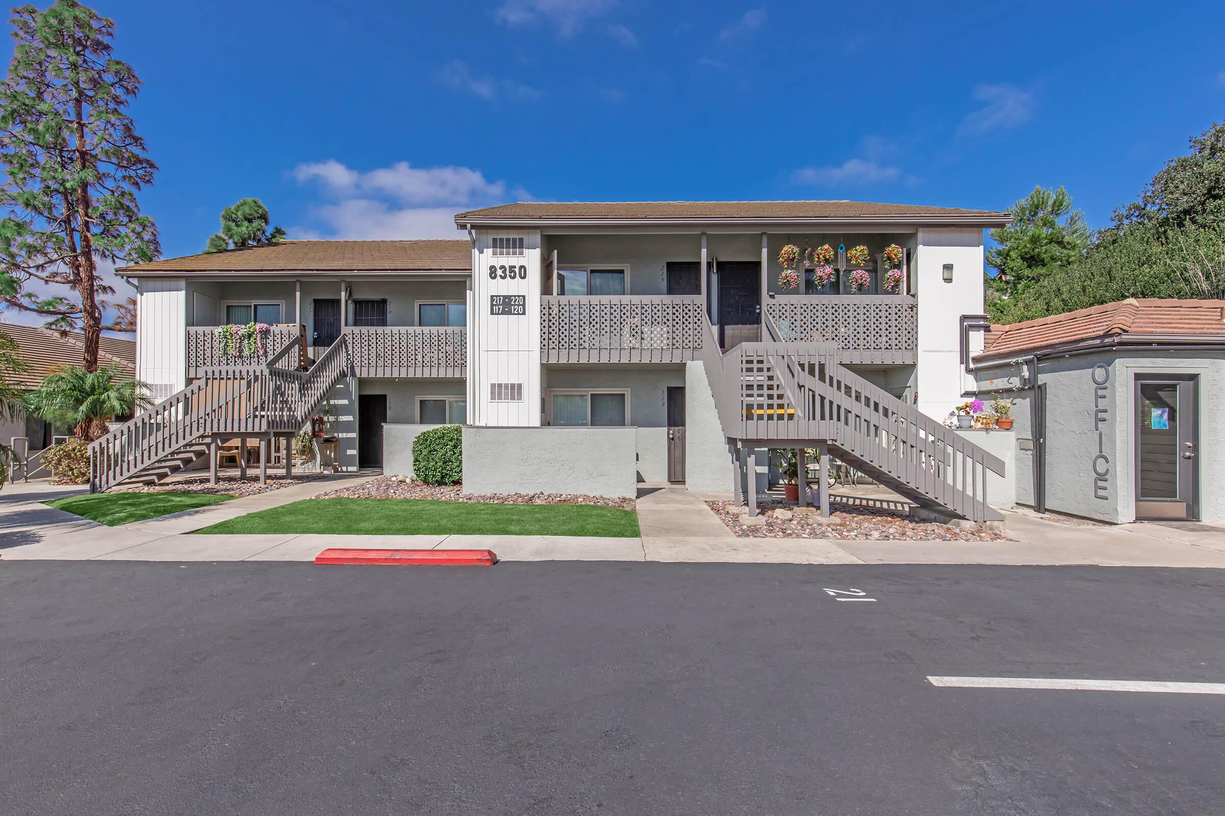 A two-story apartment building with stairs leading to individual units and flower boxes on the upper level. The building is surrounded by greenery, and there is an on-site office to the right. A clear blue sky can be seen in the background, and the parking area is in the foreground.