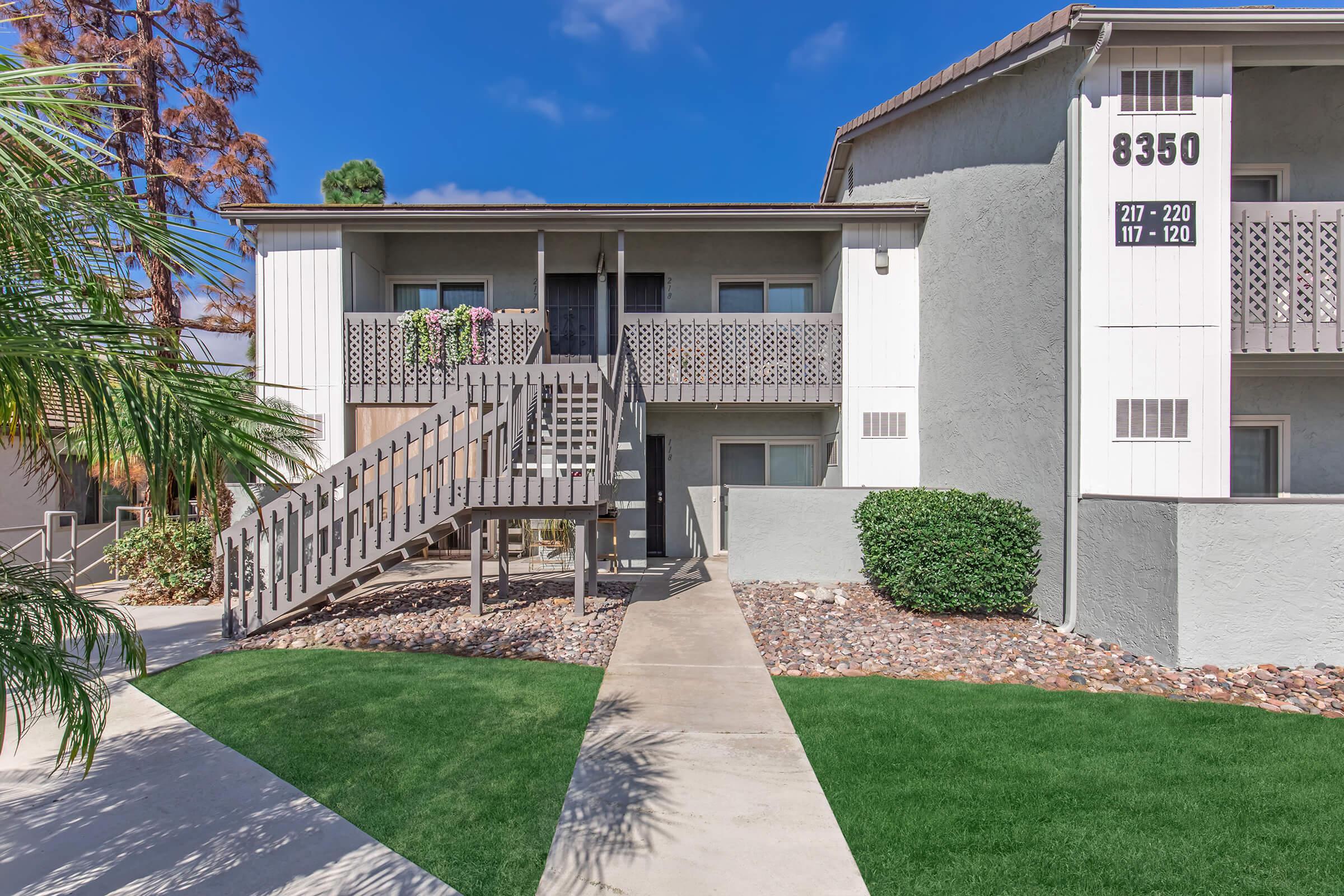 Exterior view of a multi-story apartment building featuring a staircase leading to an upper level, adorned with potted plants on the balcony. The building is surrounded by well-maintained landscaping, including green grass and decorative rocks. Clear blue sky enhances the inviting atmosphere.