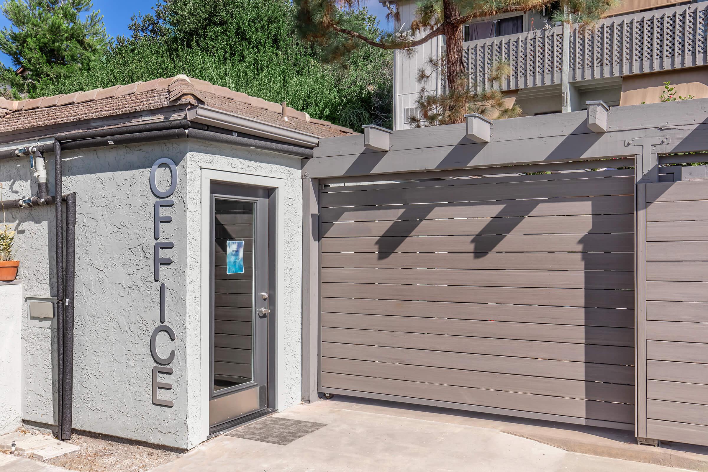A single-story building with a gray exterior features the word "OFFICE" on the wall beside a sleek door. The entrance is accompanied by a modern wooden slat gate. Surrounding greenery adds a natural touch to the setting, while the overall design conveys a contemporary aesthetic.