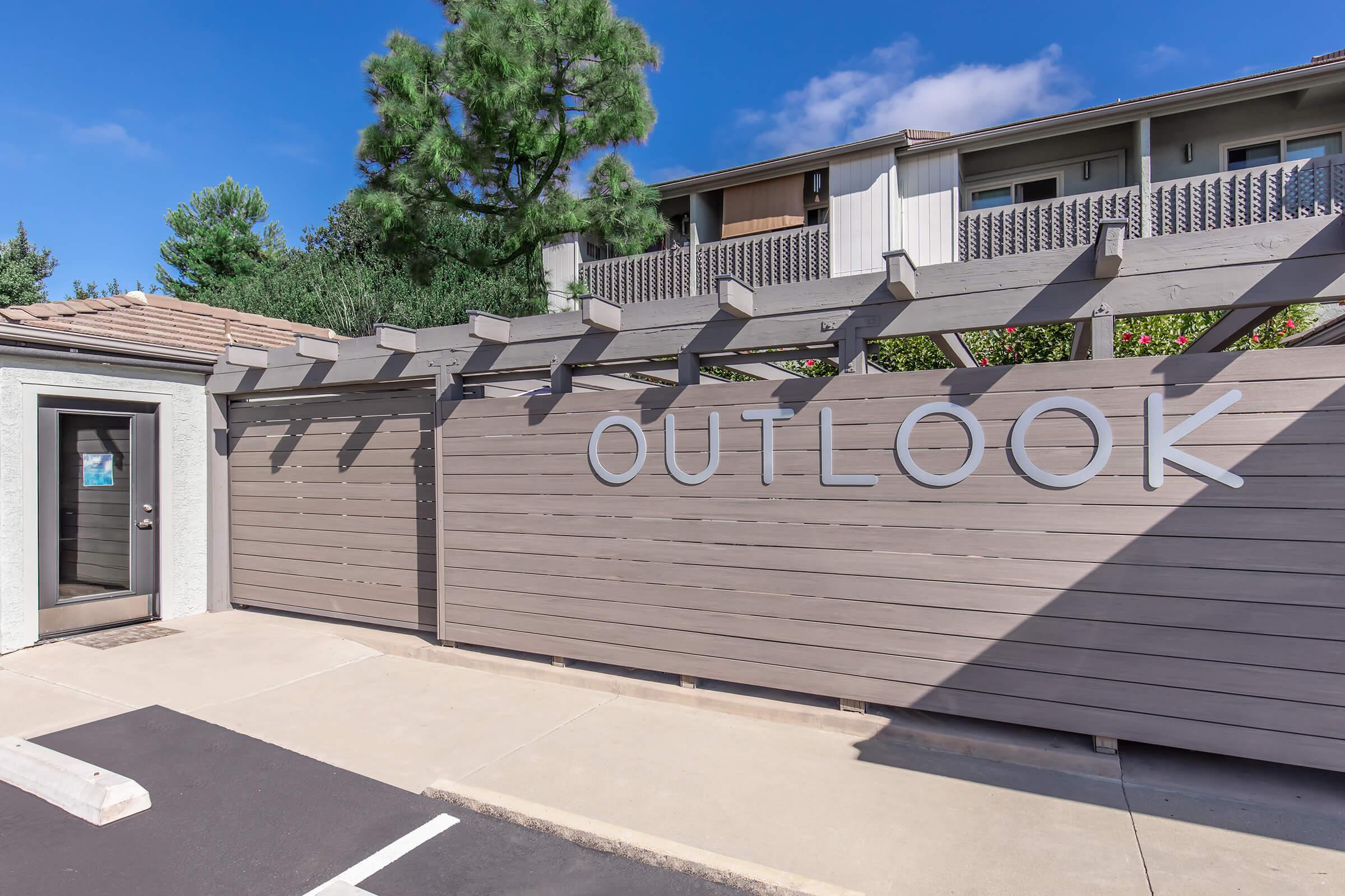 Exterior view of a building with the word "OUTLOOK" prominently displayed on a wooden wall. The entrance features a door with a simple design, and there are trees visible in the background. The area is well-maintained with a clean parking space in front.