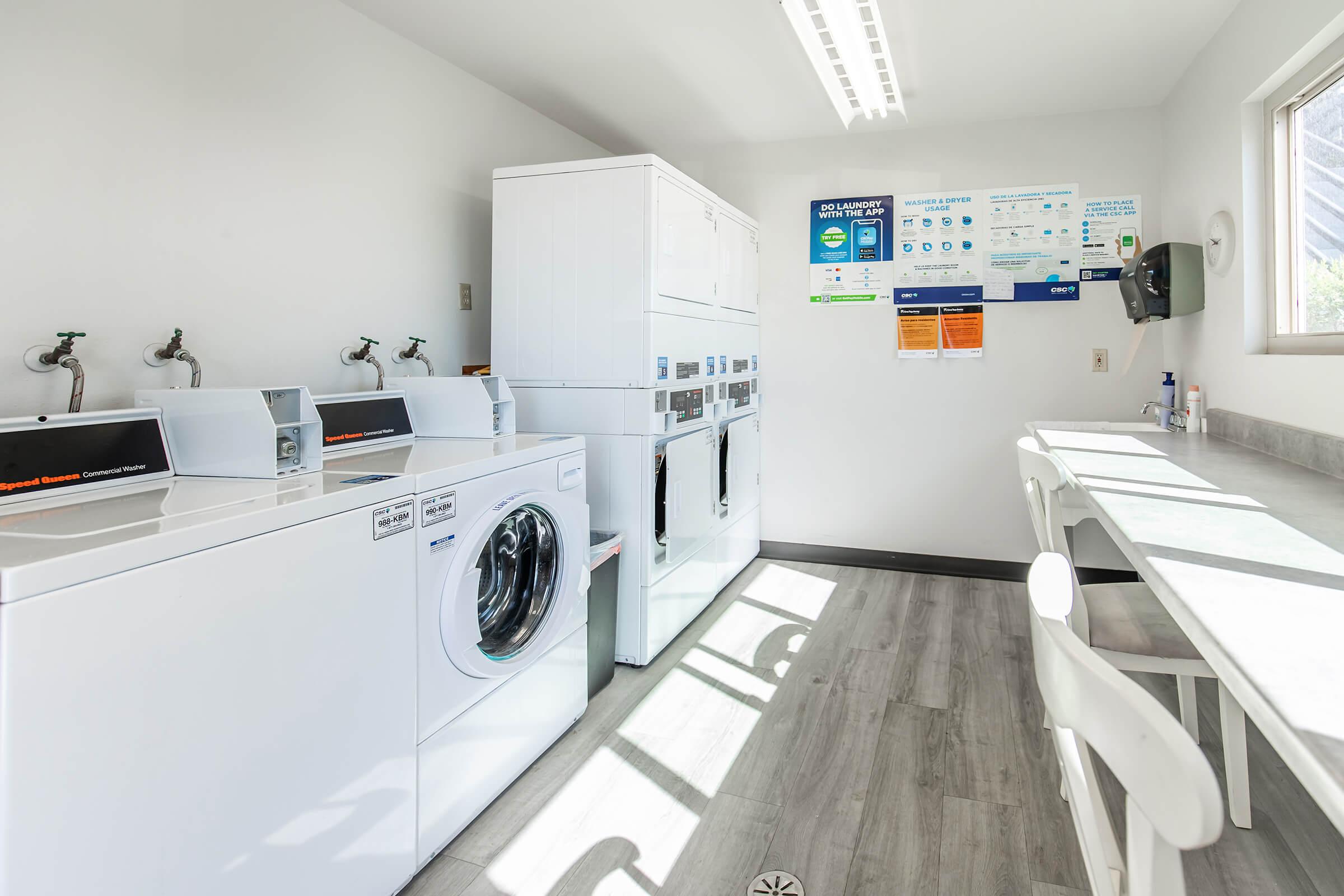 A bright, clean laundry room featuring several white washing machines and dryers. There are sinks along the wall and a table with chairs near a window. Informational posters and notices are displayed on the wall, highlighting laundry services and instructions.