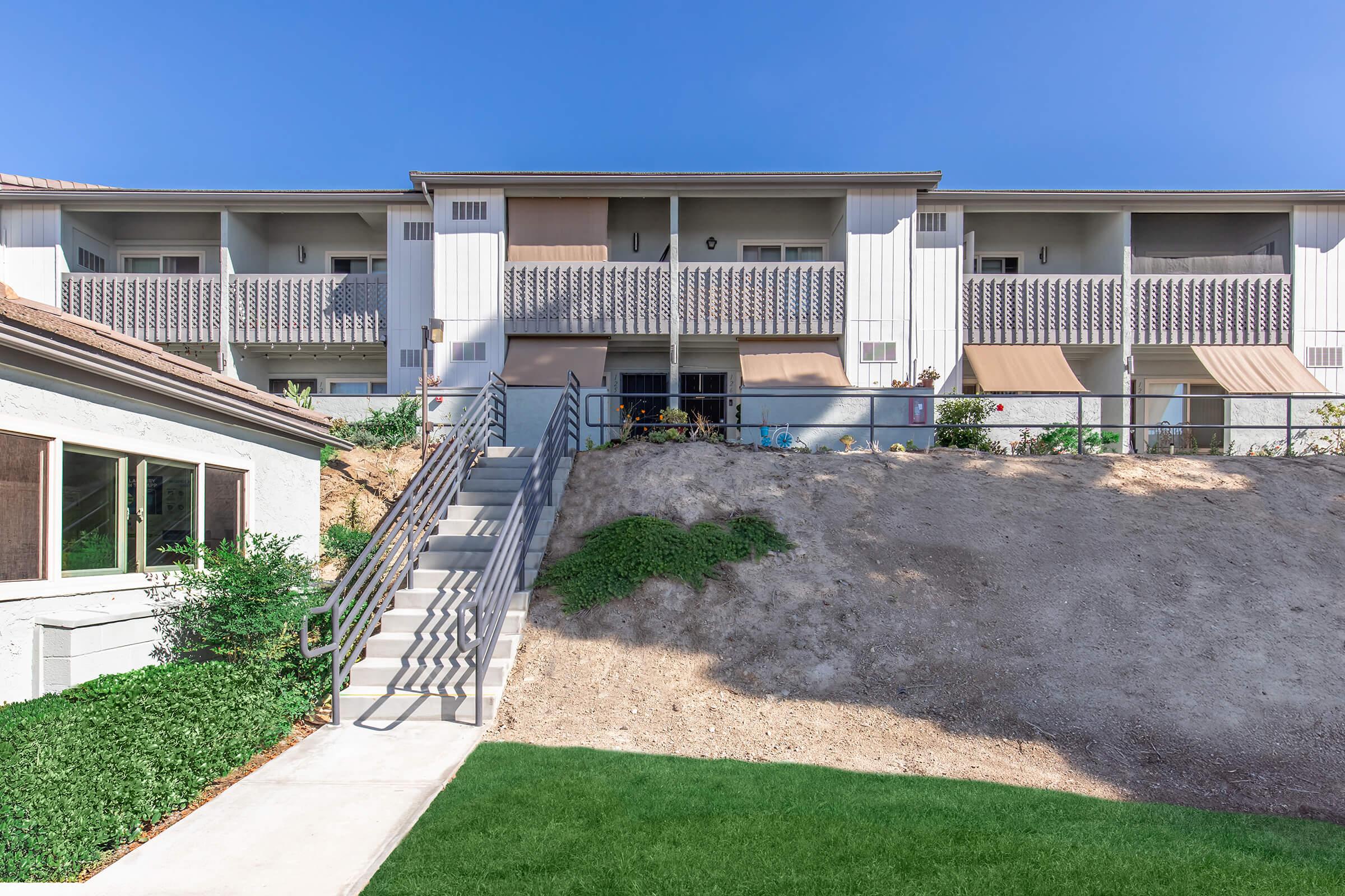 A two-story apartment building with multiple balconies, situated on a slight hillside. There is a set of stairs leading up to the entrance, and the area is landscaped with grass and shrubs. The sky is clear and blue, providing a bright backdrop to the scene.