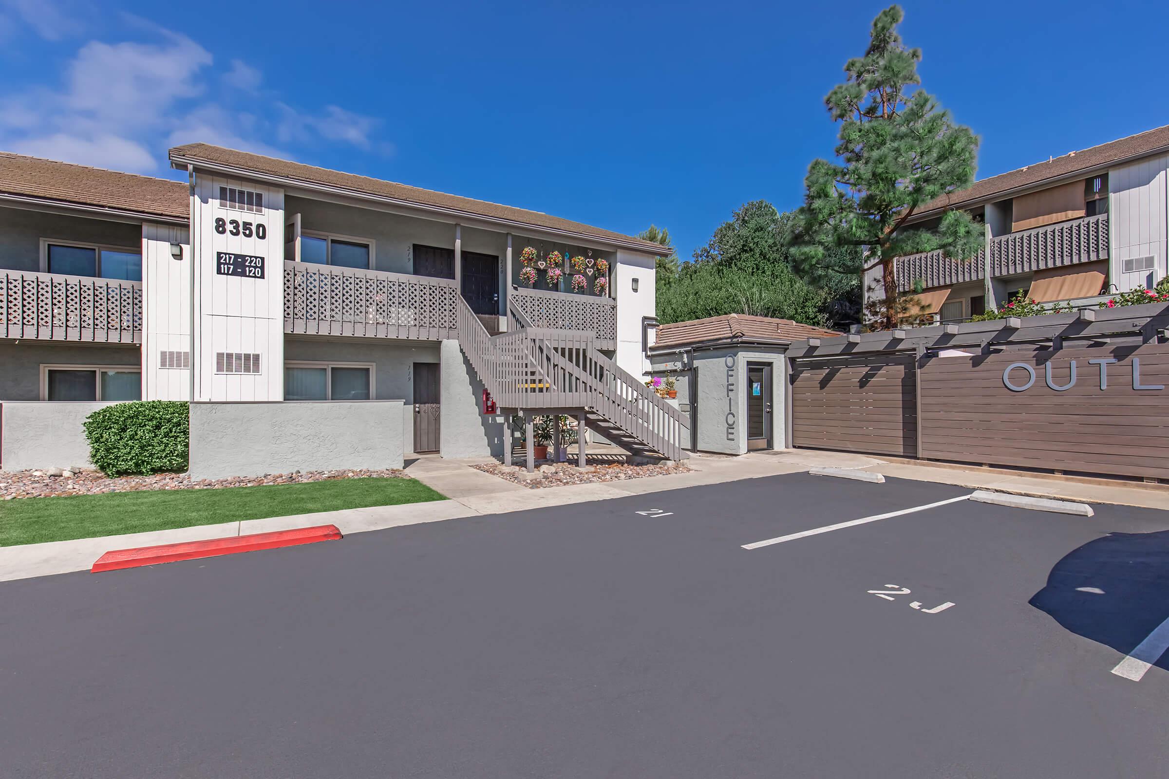 Exterior view of a multi-unit apartment building with a staircase leading to an upper level. The building features a sign with the number 8350, landscaped areas with shrubs, and a parking lot with marked spaces. Clear blue skies and a few trees are visible in the background.