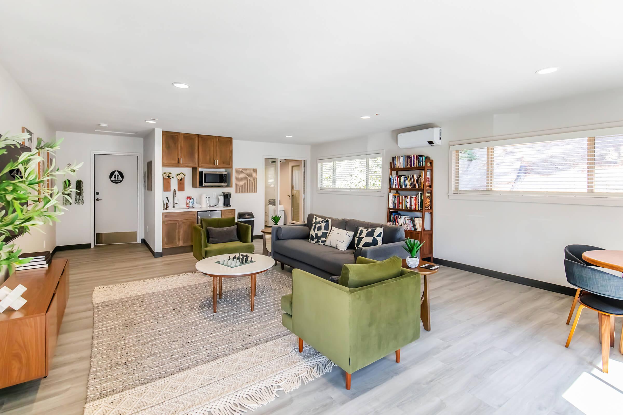 A stylish living room featuring a gray sofa, green armchairs, and a round coffee table on a light rug. The space is bright with natural light from large windows, showcasing a modern kitchen area and a bookshelf in the background. The decor is contemporary and inviting, creating a cozy atmosphere.