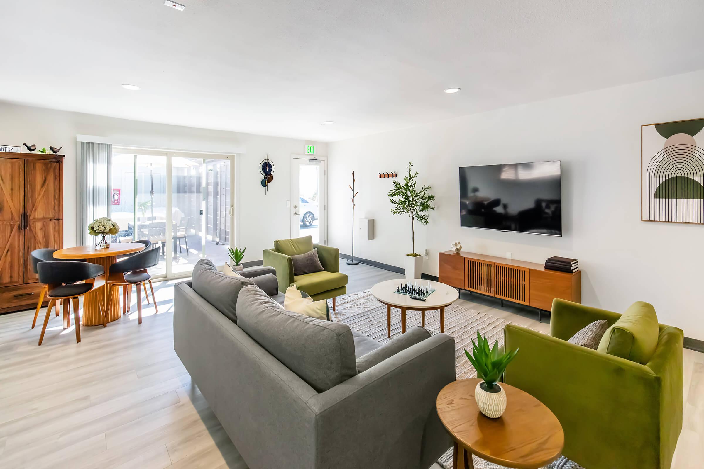 A modern living room featuring two gray sofas and two green armchairs, a round coffee table with a plant, and a large wooden TV console. A dining table is visible in the background, along with a sliding glass door leading to an outdoor area. Minimalistic decor accents the bright, inviting space.