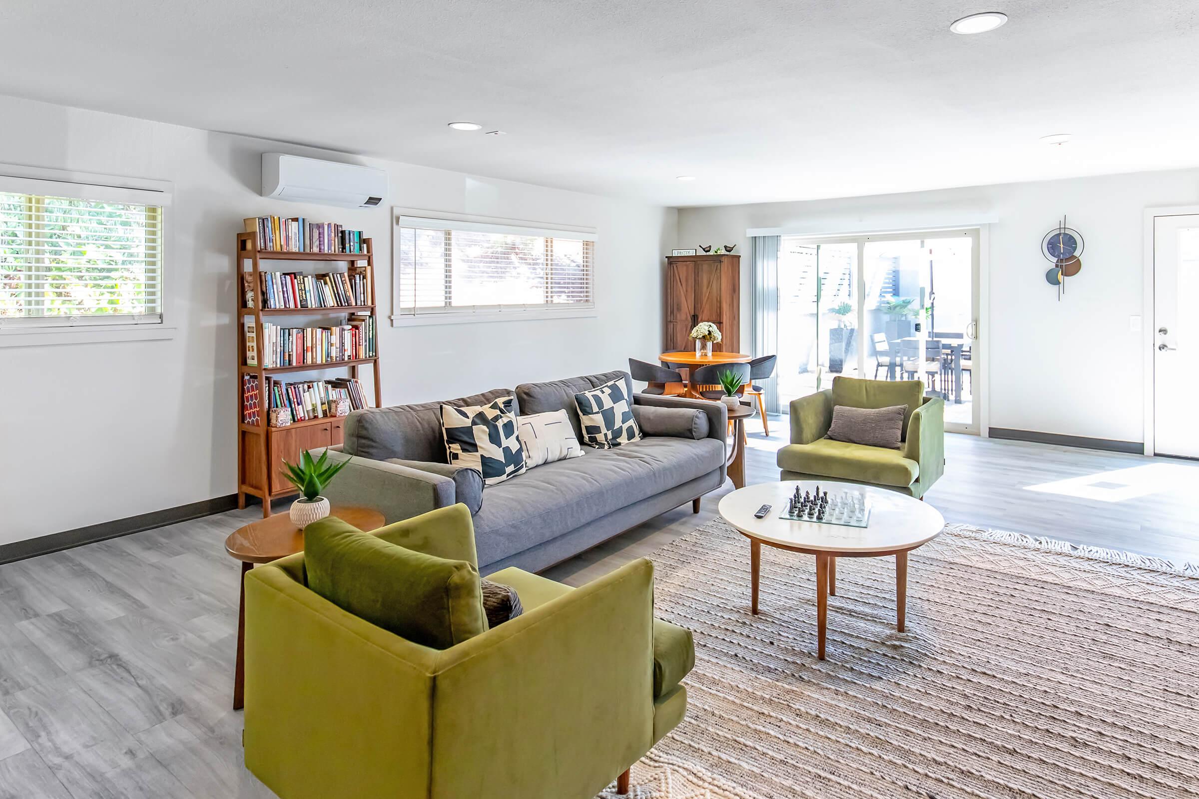 A modern living room featuring a gray couch with decorative pillows, two green armchairs, a round wooden coffee table with a chess set, and a bookshelf filled with books. Large windows allow natural light, and a door leads to an outdoor patio area. The floor is light gray, enhancing the bright and airy feel of the space.