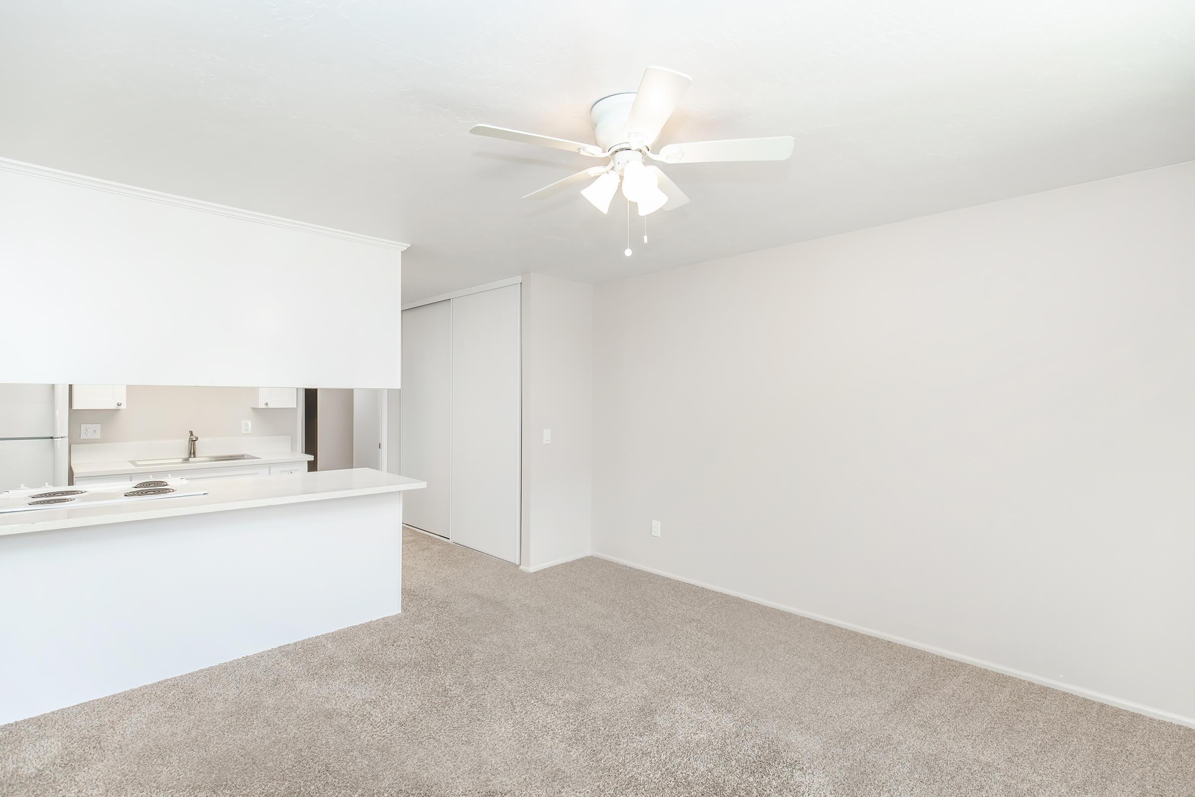 A spacious, well-lit living area with light-colored walls and carpet. There is a ceiling fan and a small kitchen area in the background, partially visible. The space appears clean and ready for furniture arrangements, with a sliding door closet on one side.