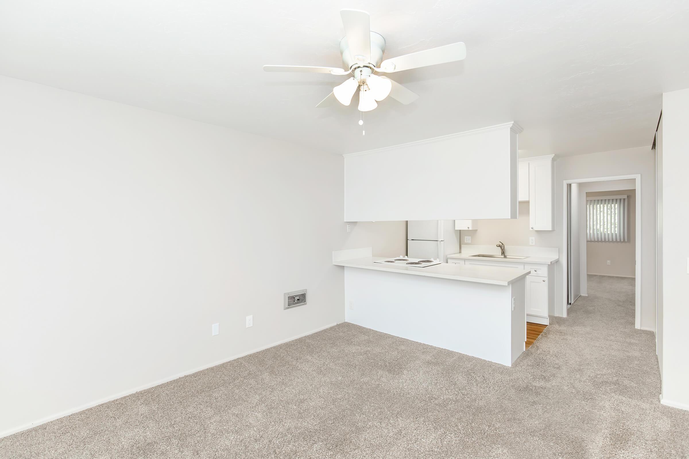 Bright and airy living space featuring a ceiling fan, beige carpet flooring, and a kitchenette with white cabinetry. The open layout allows for natural light, with a small window visible in the background. The walls are painted in a light color, creating a welcoming atmosphere.