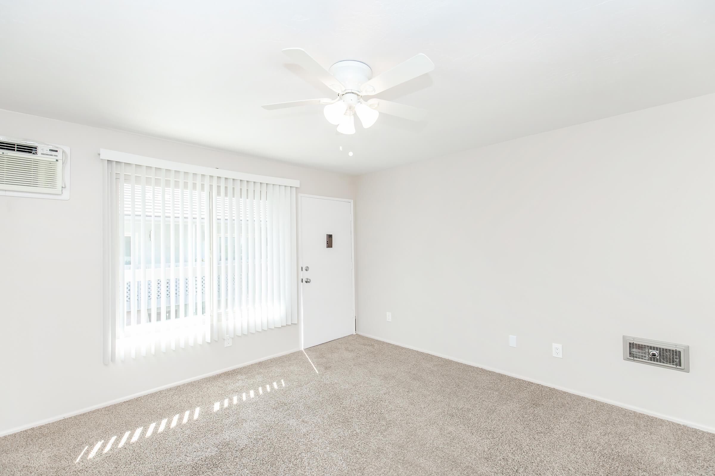A bright and empty room featuring light-colored walls and beige carpet. A ceiling fan is installed, and there are vertical blinds on the windows. A door is visible, and there's an air conditioning unit on one wall, creating a spacious and airy atmosphere.
