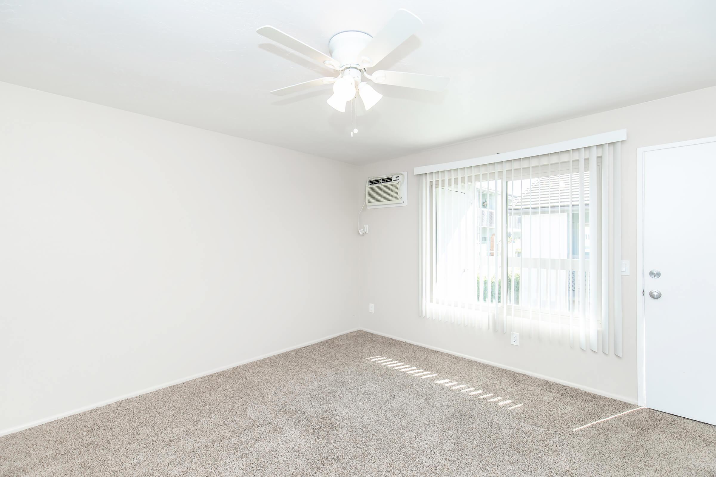 A bright, empty room with light beige walls and carpet. There is a ceiling fan with white blades and an air conditioning unit on the wall. Vertical blinds cover a window that lets in natural light. A white door is visible on one side of the room, enhancing the spacious feel.