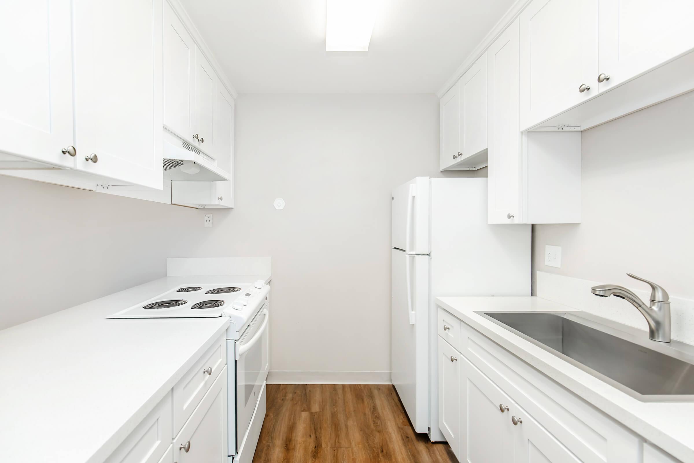 Modern kitchen featuring white cabinetry, a white stove, and a refrigerator. The countertops are light-colored, and there is a stainless steel sink with a sleek faucet. The flooring is a warm wood finish, and the walls are painted in a neutral tone. The space is well-lit with overhead lighting.