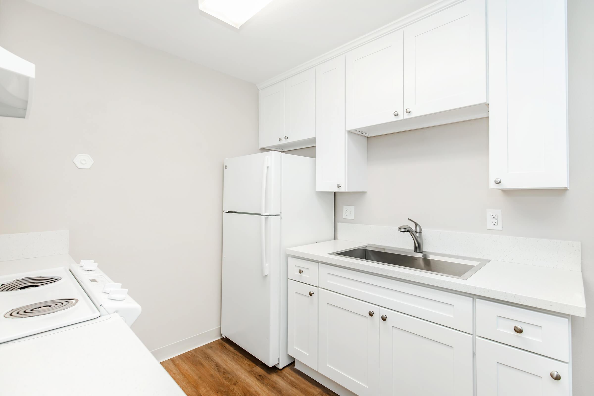 A modern kitchen featuring white cabinets, a stainless steel sink, and a white refrigerator. The countertop is light-colored and the flooring is wood. There’s a stove with an oven on the left, and the walls are painted in a neutral shade, creating a bright and clean space.