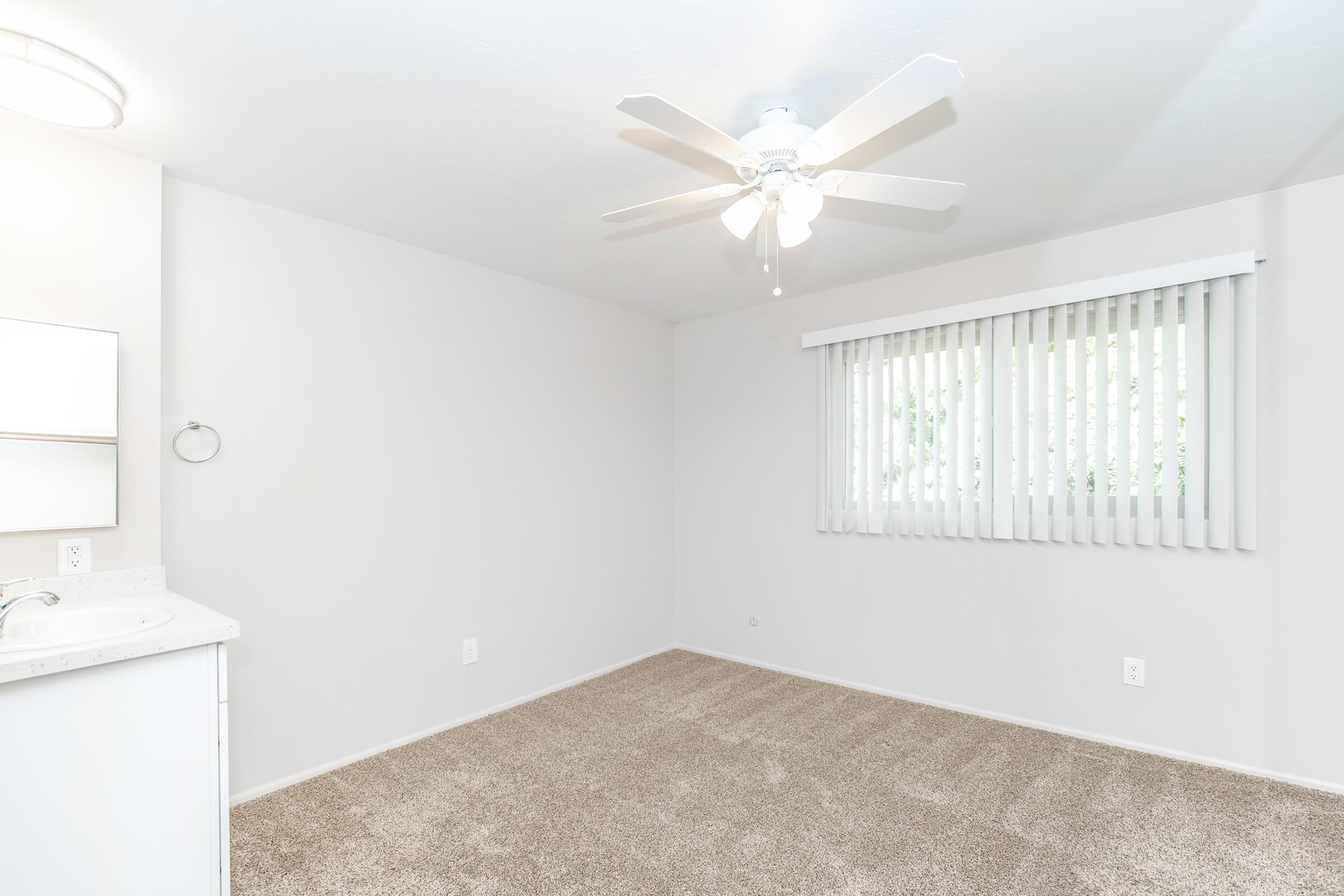 Empty room featuring light gray walls and beige carpet. A ceiling fan with light fixture is mounted above, and a window with blinds allows natural light. A small, white countertop with a sink is visible on the left side, and a towel holder is mounted on the wall nearby. The space is bright and uncluttered.
