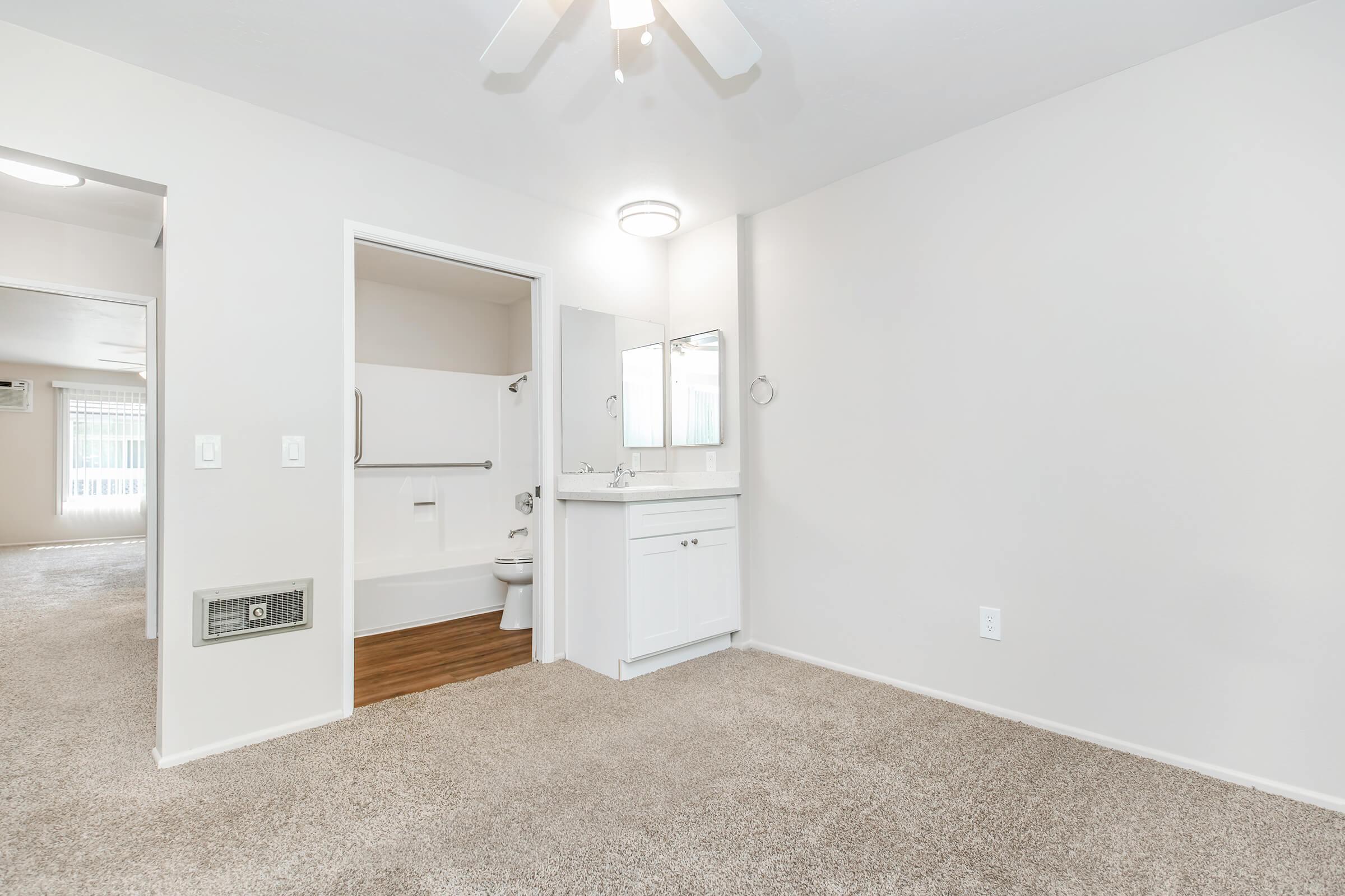 A well-lit, vacant room featuring beige carpet and neutral-colored walls. There is a small bathroom area on the left with a sink, mirror, and a toilet. A ceiling fan is visible, and a doorway leads to another space. The overall ambiance is clean and inviting, suitable for various uses.