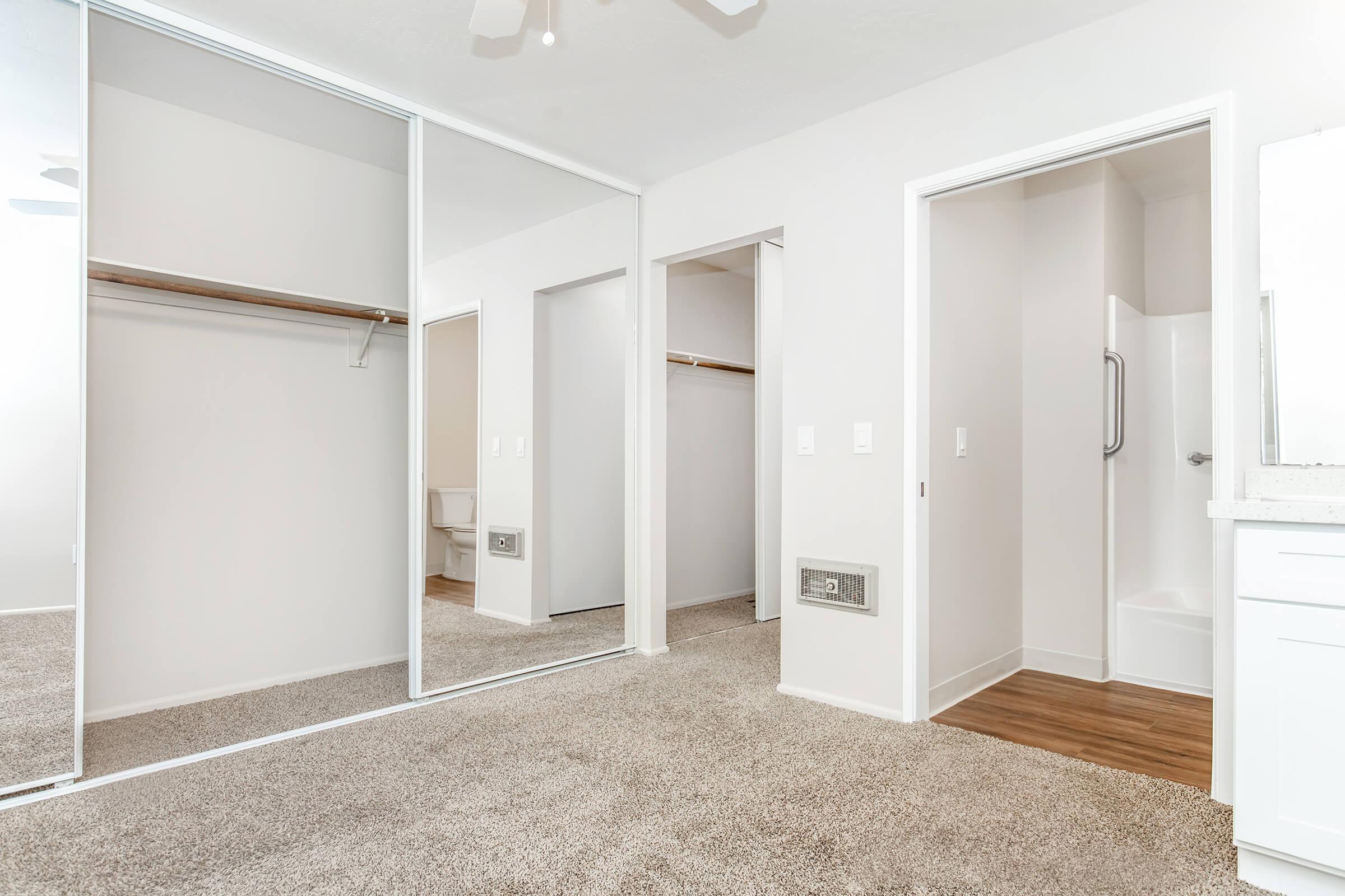 A clean, well-lit bedroom featuring two mirrored closet doors, a small bathroom entrance with a shower, and plush carpet flooring. The walls are painted in a neutral color, and the overall aesthetic is modern and minimalist.