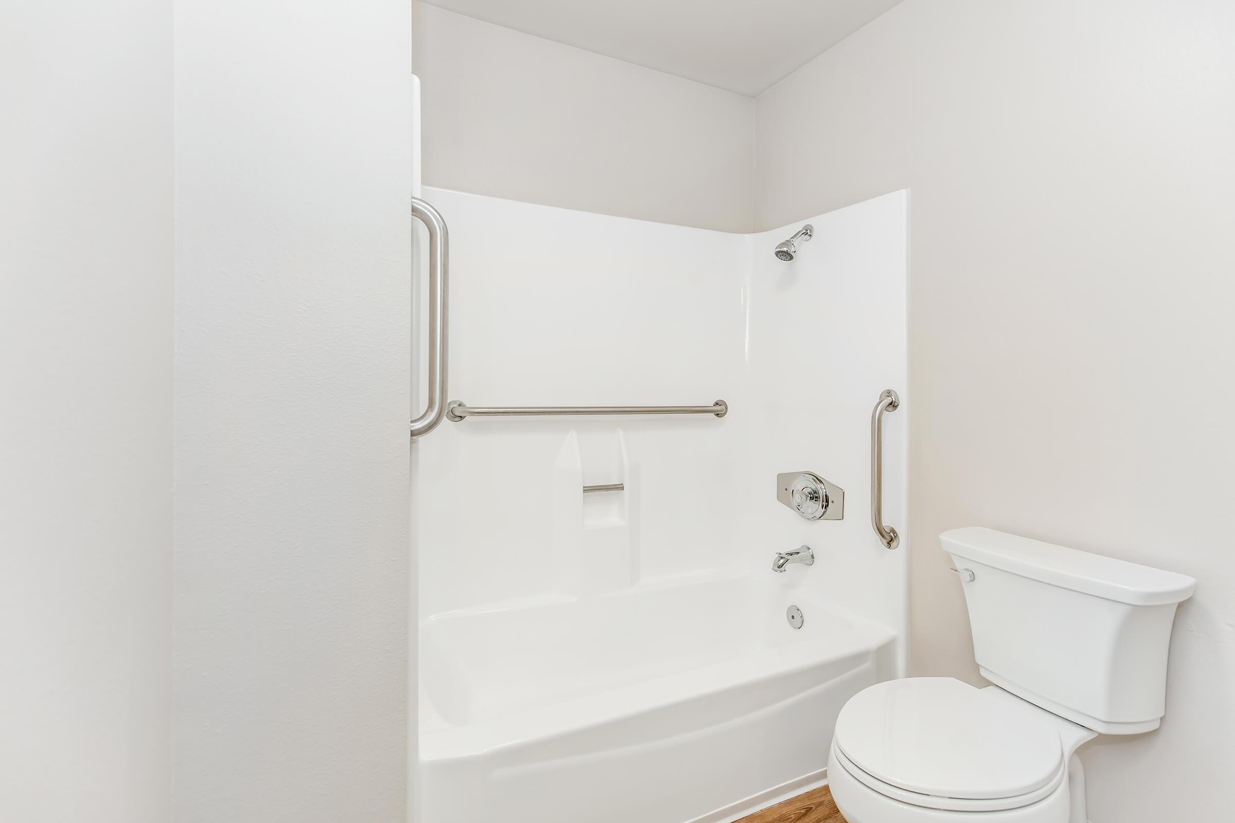 A clean, modern bathroom featuring a white bathtub with a showerhead, grab bars for safety, and a white toilet. The walls are light-colored, and the flooring appears to be wood or laminate. The space is well-lit and designed for accessibility.