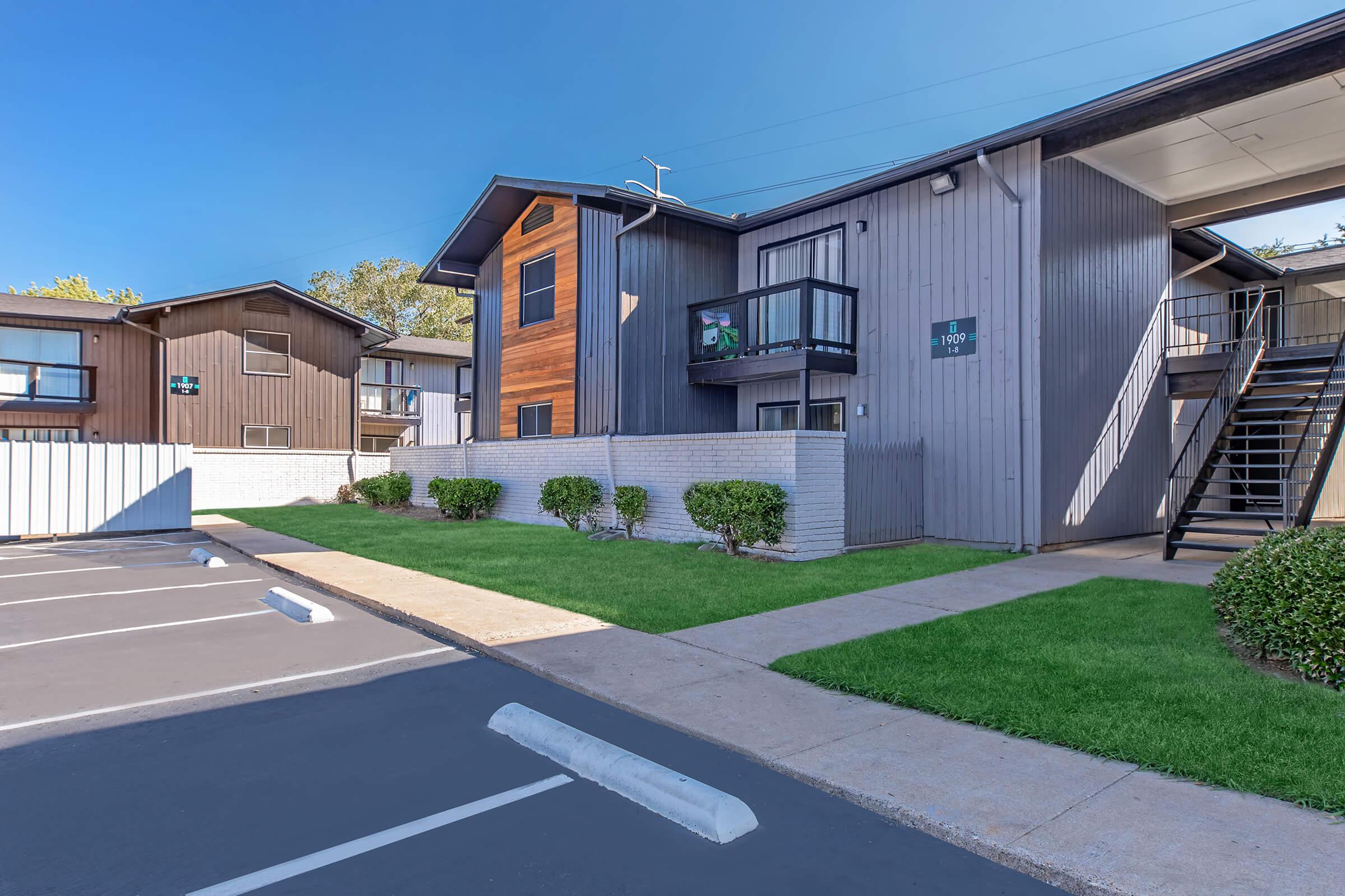 A modern apartment complex featuring two-story buildings with a mix of wooden and gray exterior finishes. Well-maintained landscaping with neatly trimmed bushes and a paved pathway leading to the entrance. Parking spaces are visible in the foreground, with clear blue skies above.