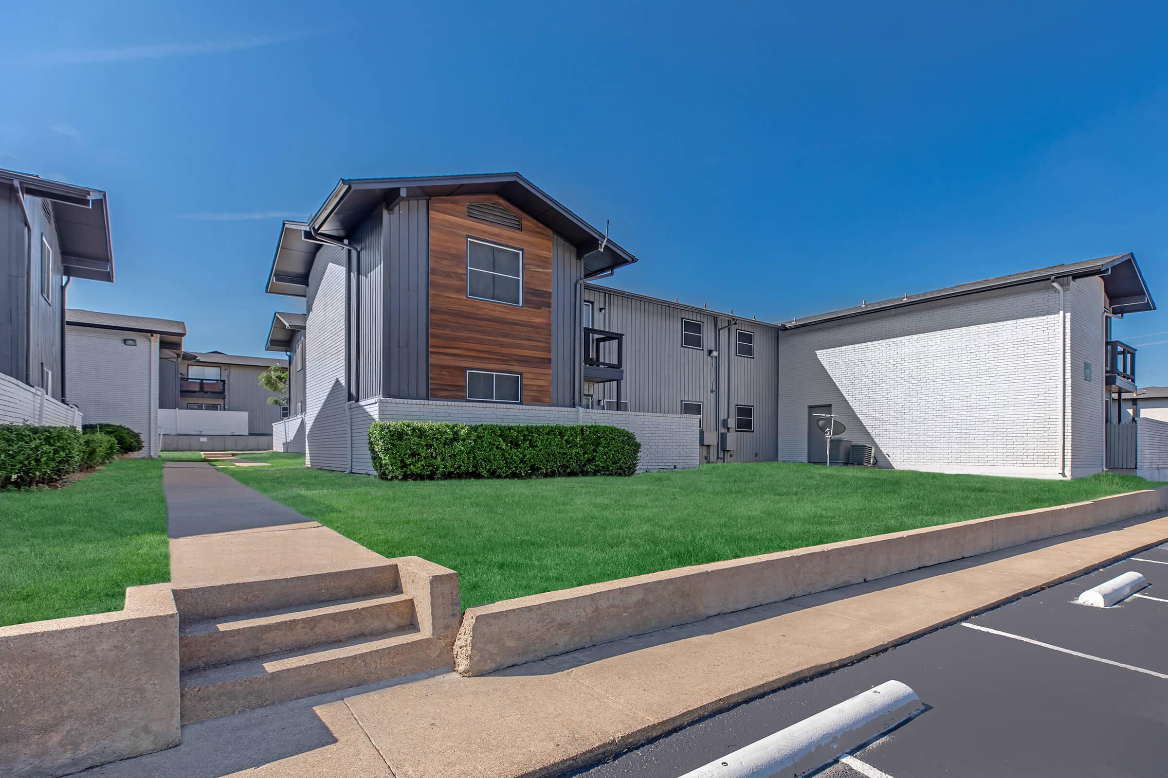 Modern apartment complex with a mix of gray and brown siding, featuring multiple units. There is a well-maintained green lawn in front, with a walkway leading to the entrance. The blue sky above indicates clear weather, and parked cars are visible along the driveway.