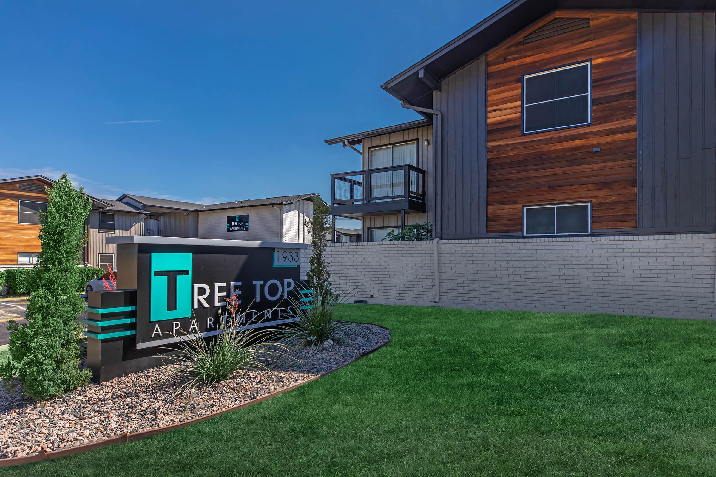 Sign for Tree Top Apartments with the building in the background. The sign features a modern design with green and blue accents and is surrounded by well-maintained landscaping, including grass and shrubs. The apartment buildings have a mix of wooden and painted exteriors under a clear blue sky.