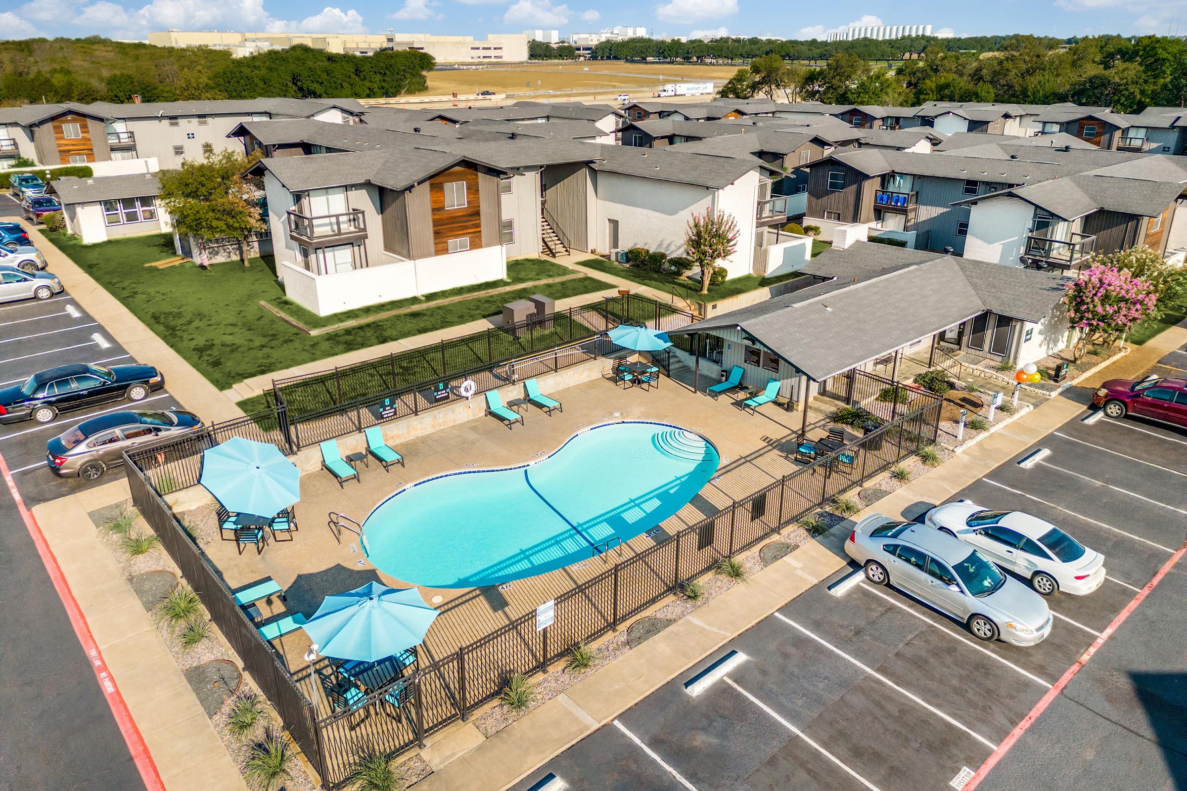Aerial view of an apartment complex featuring a swimming pool surrounded by lounge chairs and umbrellas. Well-maintained landscaping and parking spaces for cars are visible, along with several two-story buildings in the background under a clear sky.