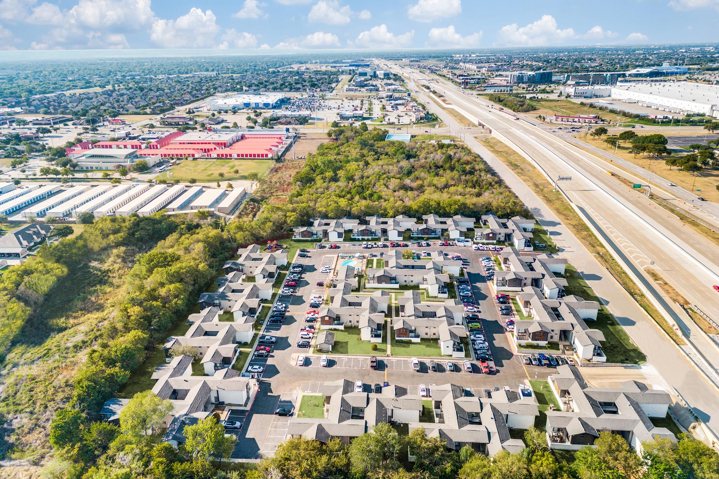 Aerial view of a residential complex featuring multiple single-story buildings arranged around a central green space. Surrounding areas include industrial and commercial properties, as well as a highway. The scene is set against a clear blue sky with scattered clouds.