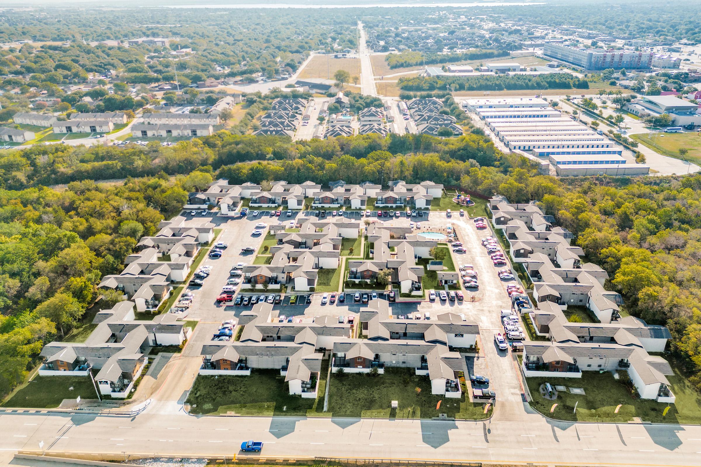 Aerial view of a residential neighborhood featuring multiple two-story houses arranged in a circular layout. Surrounding the area are trees and open fields, with additional buildings visible in the background, possibly commercial or industrial. Roads are seen connecting the houses, and parked cars are scattered throughout.