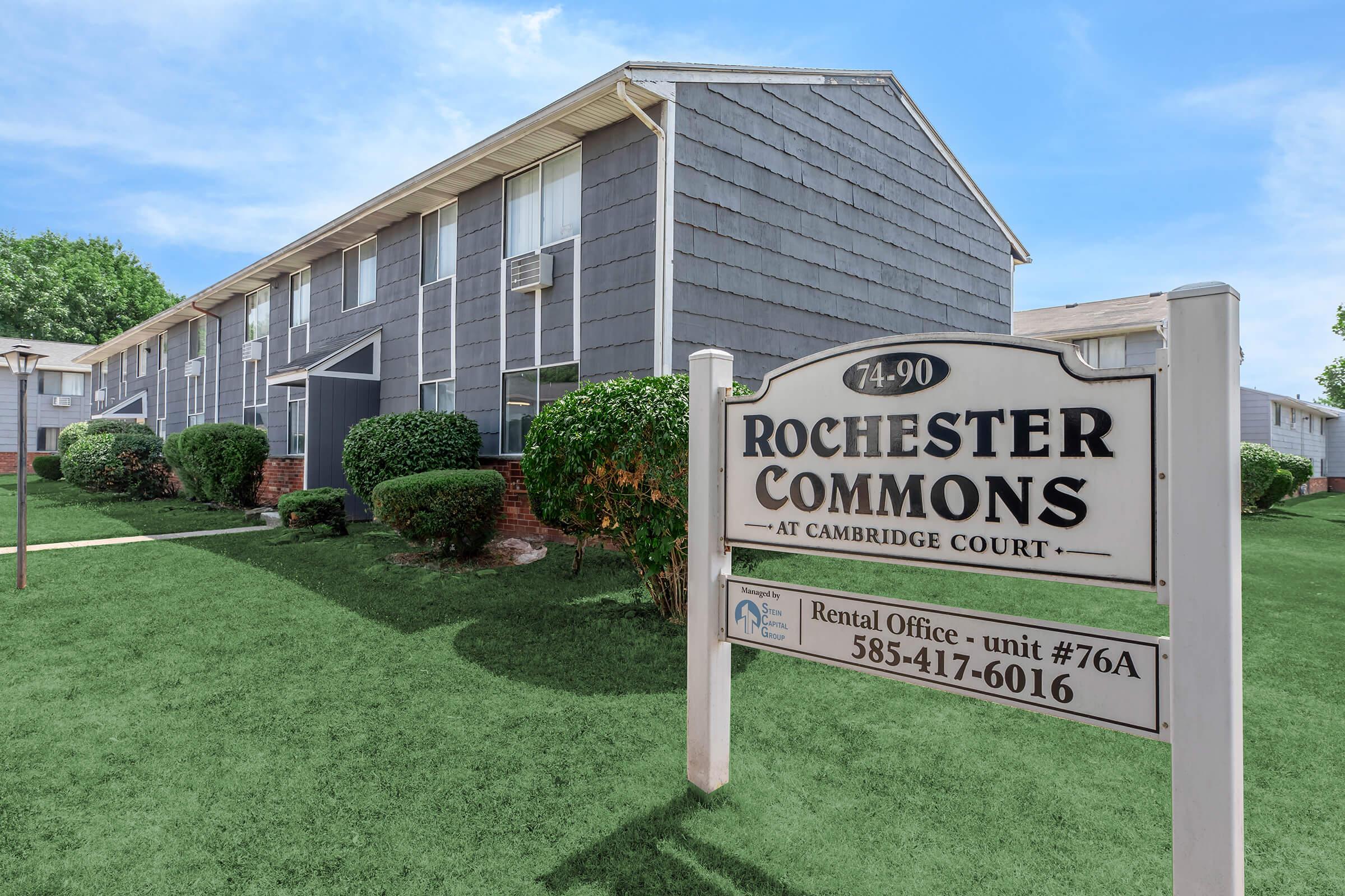 Exterior view of Rochester Commons at Cambridge Court, featuring a two-story apartment building with grey siding, large windows, and manicured green lawns. A sign prominently displays the name "Rochester Commons" along with the address and contact information for the rental office.