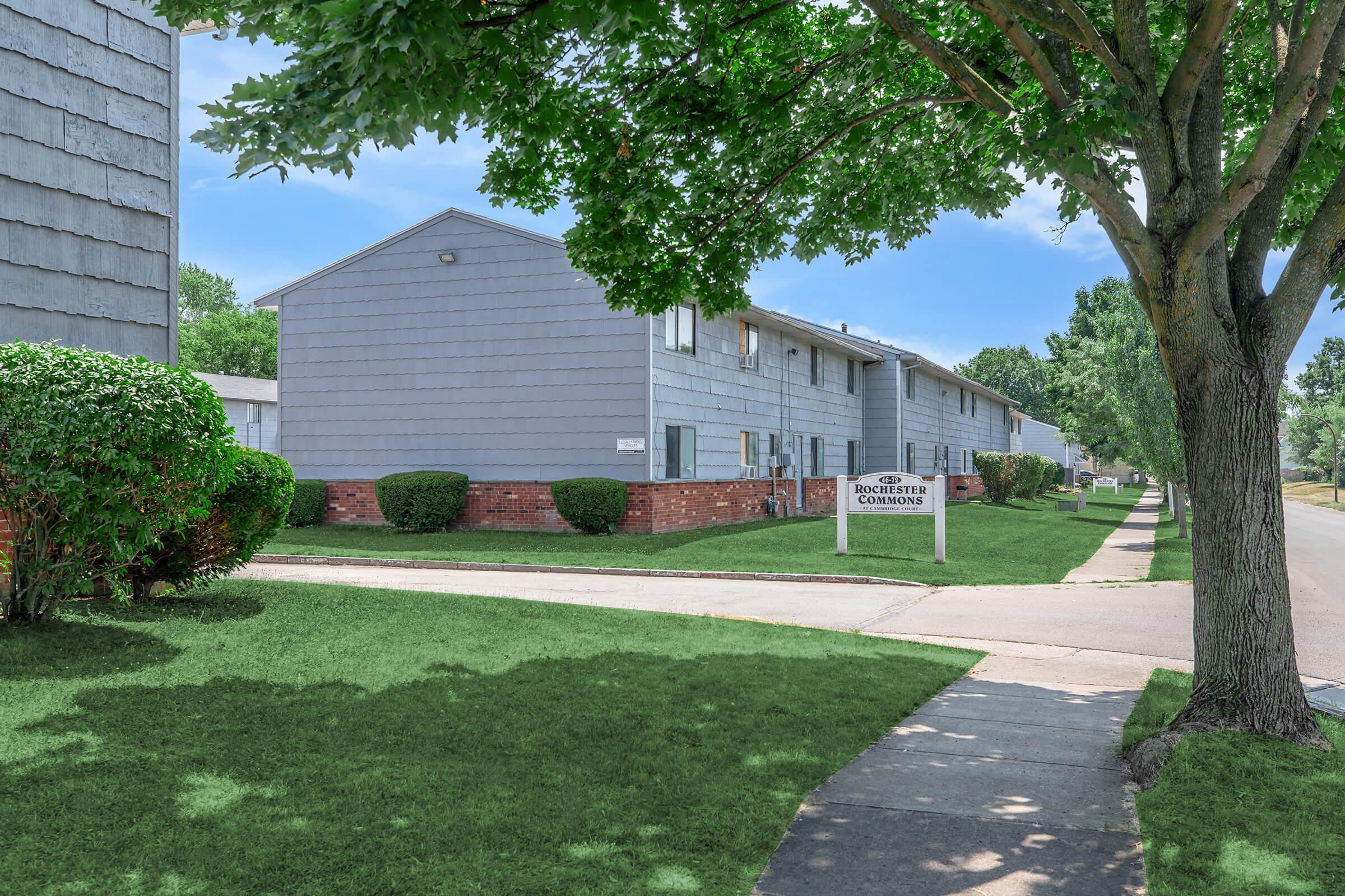 A view of a suburban apartment complex featuring gray two-story buildings surrounded by green lawns and neatly trimmed bushes. A sidewalk runs alongside the buildings, leading to a sign that reads "Edgewater Commons." The scene is bright with clear skies and well-maintained landscaping.