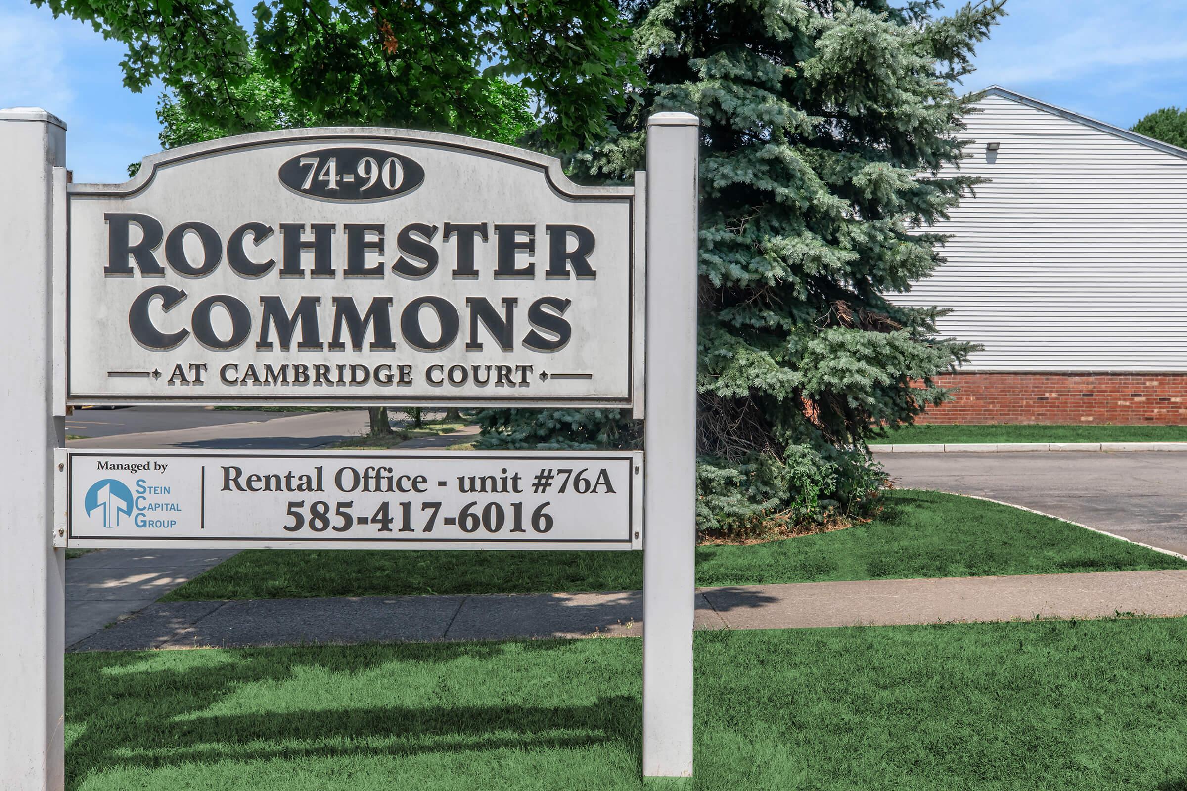 Sign for Rochester Commons at Cambridge Court, displaying the address 74-90, along with information about the rental office located in unit #76A. Contact number provided is 585-417-6016, and it indicates management by Stein Capital Group. Surrounding greenery and a residential building are visible nearby.
