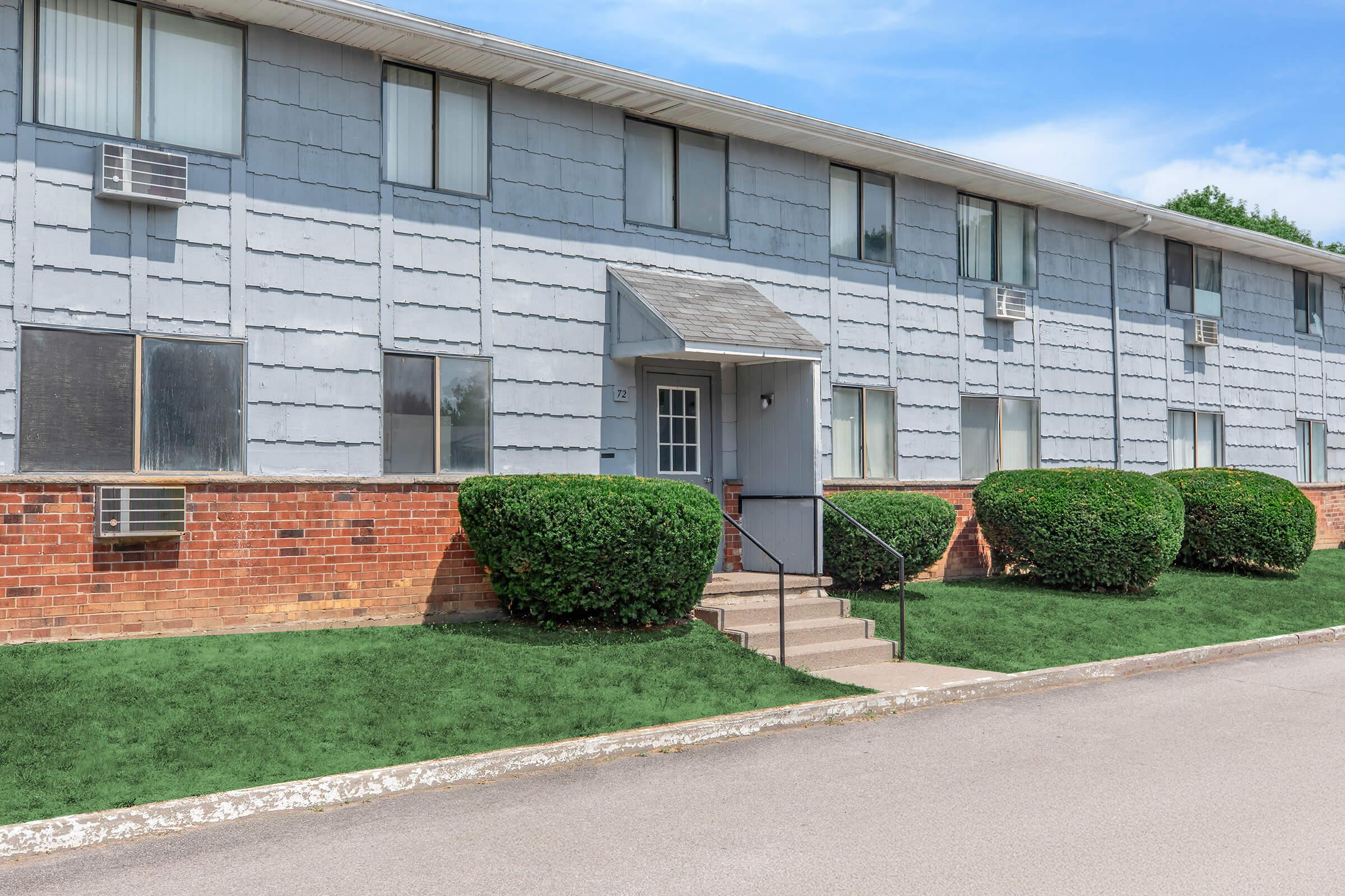Exterior view of a two-story apartment building with a gray and brick facade. The building features multiple air conditioning units and a small set of stairs leading to the entrance. Well-maintained shrubs and grass surround the building, under a clear blue sky.