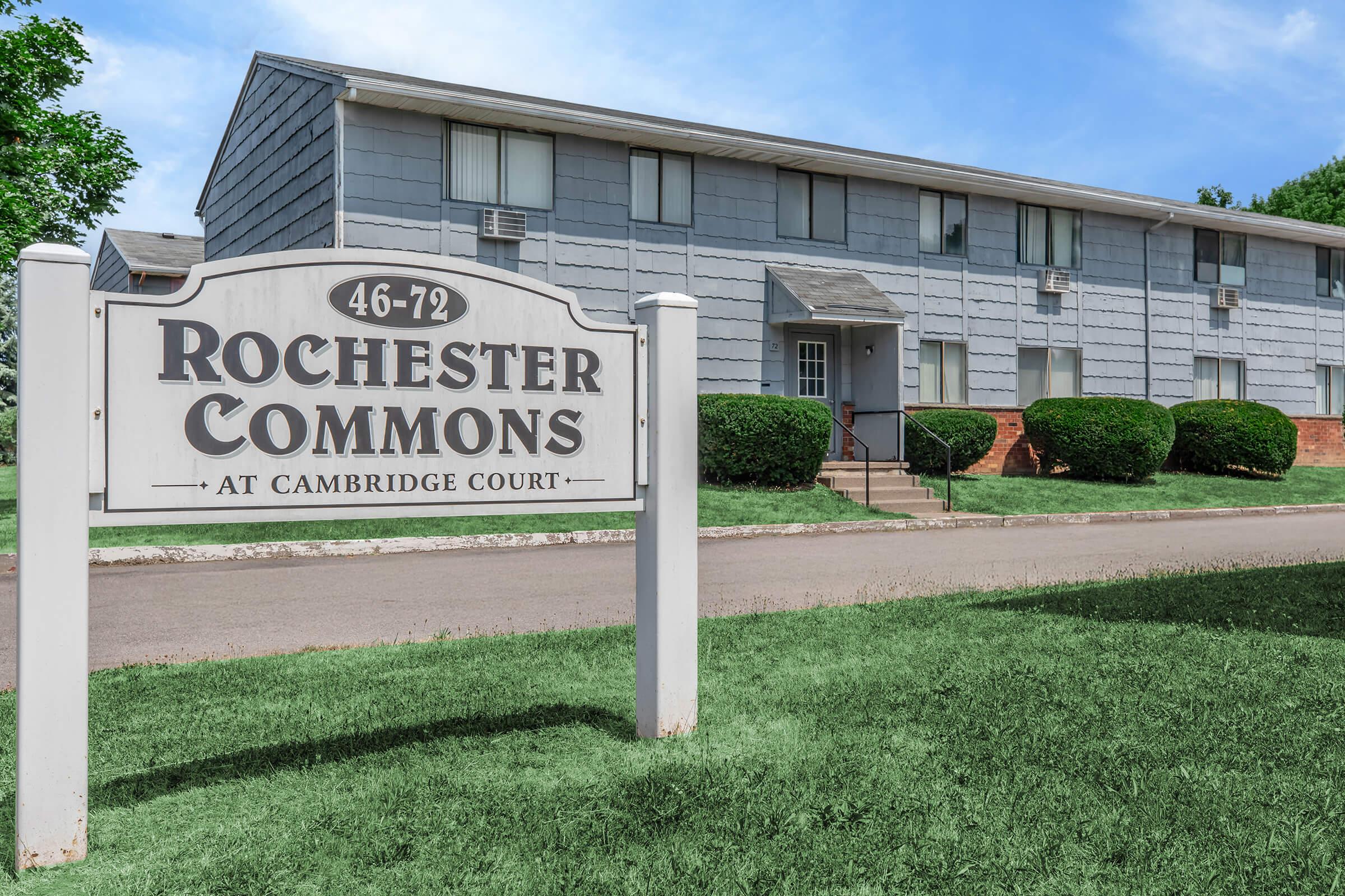Sign for Rochester Commons at Cambridge Court in front of a multi-unit residential building. The building features gray siding, multiple windows, and a walkway leading to the entrance. The surrounding area has well-maintained grass and shrubs.