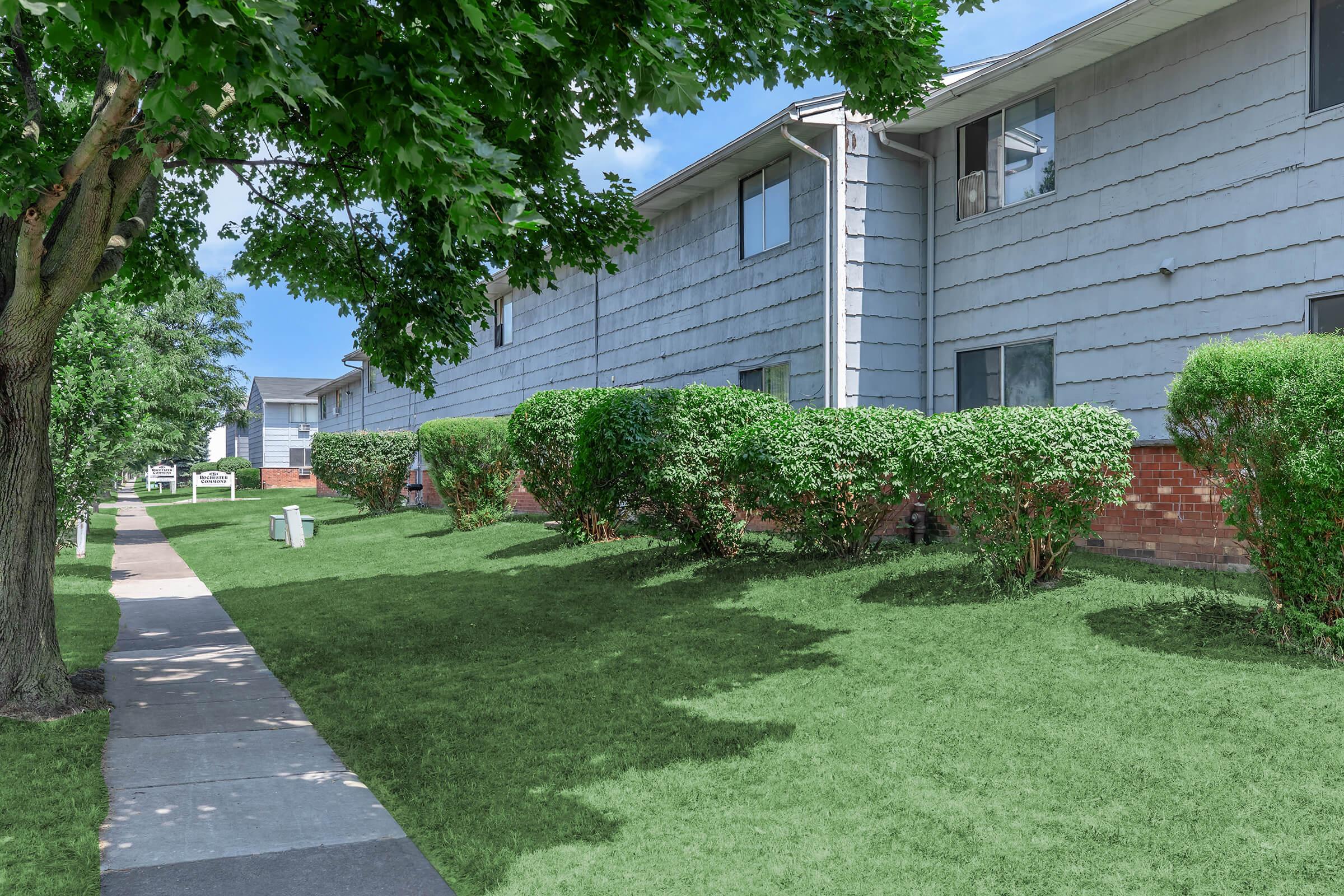 A pathway lined with lush green grass and shrubs leads to a row of residential buildings. The buildings have a light gray exterior with multiple windows. Tall trees provide shade along the sidewalk, creating a pleasant and inviting neighborhood atmosphere on a sunny day.