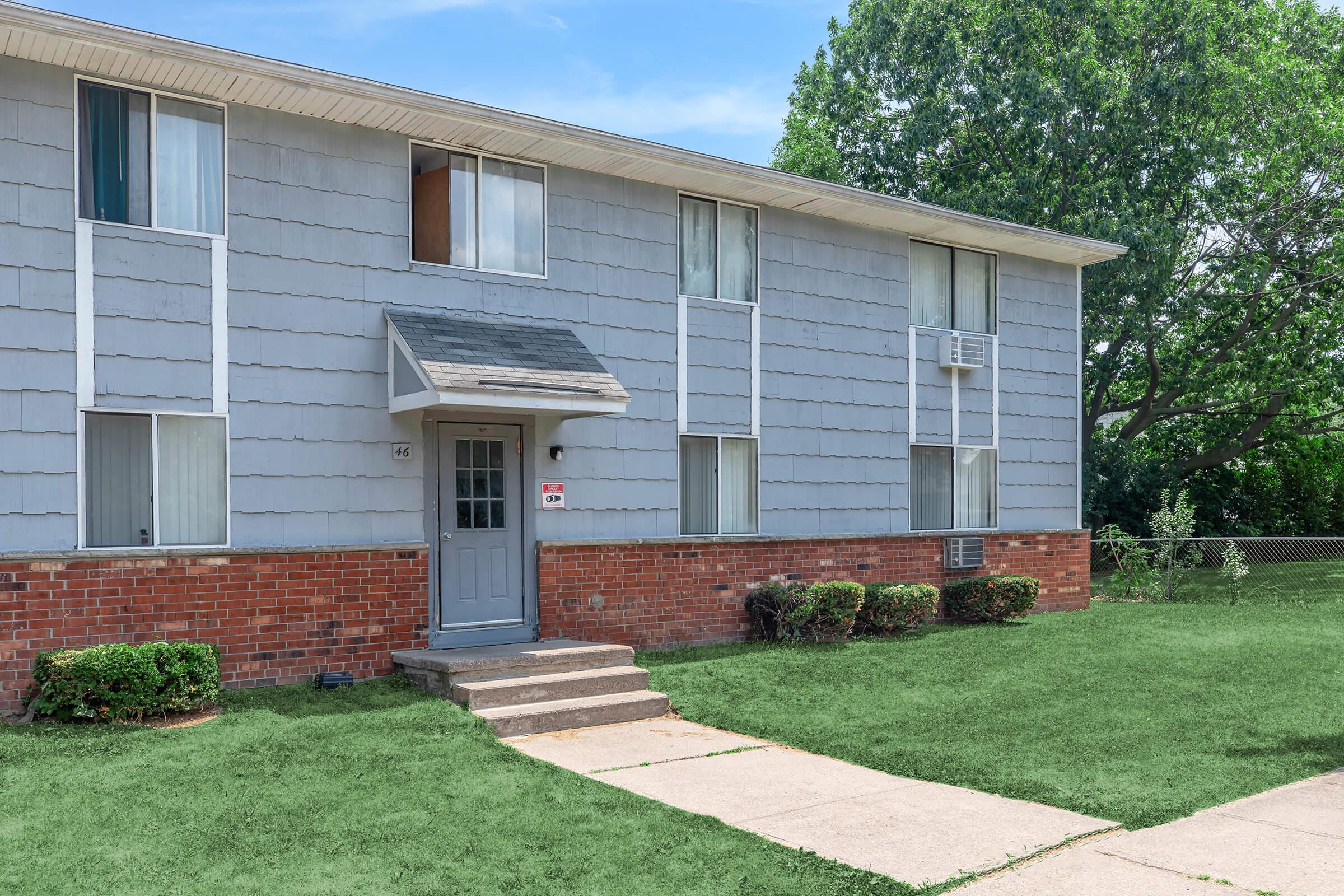 A two-story apartment building with gray siding and a brick base. The entrance features a small covered porch with a gray door. The surrounding area has green grass and neatly trimmed bushes. A sidewalk leads up to the entrance, and the building is set against a clear blue sky with trees in the background.