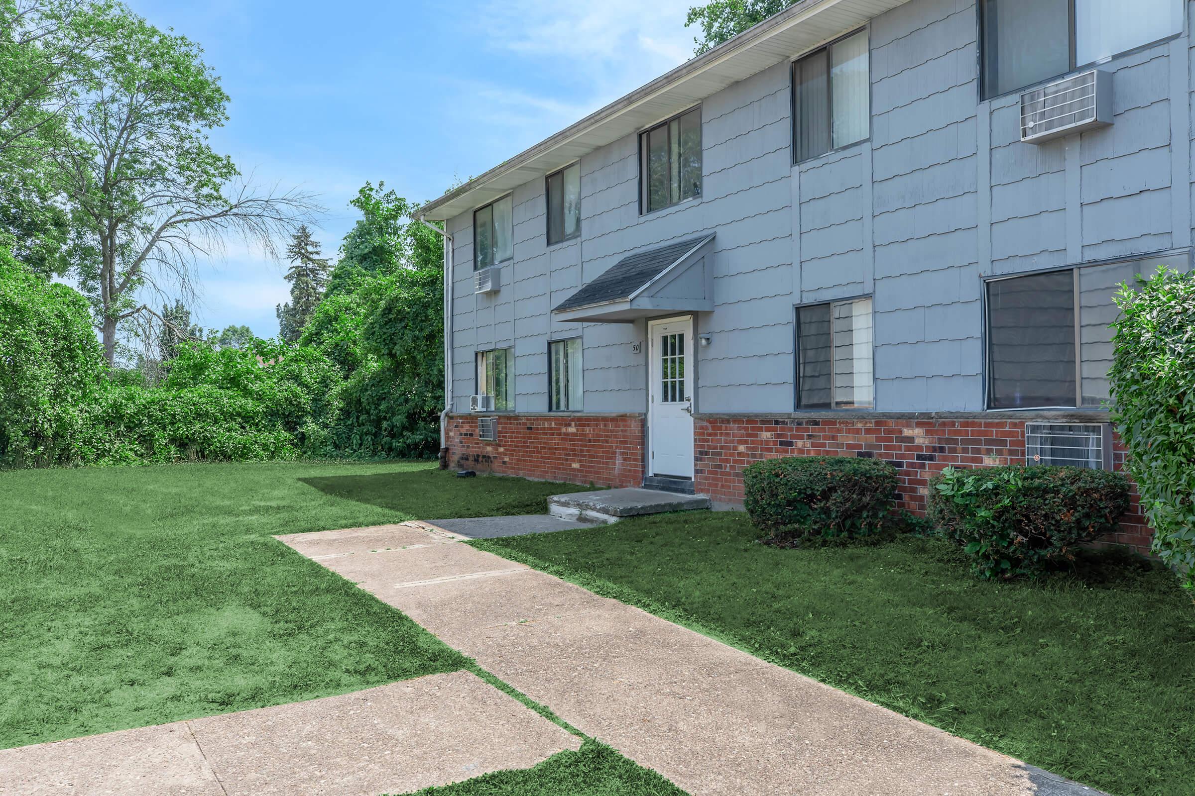 A two-story residential building with a gray exterior and brick accents, surrounded by green grass and shrubs. There are multiple windows with air conditioning units, and a concrete pathway leading to the front door. Trees are visible in the background under a clear blue sky.