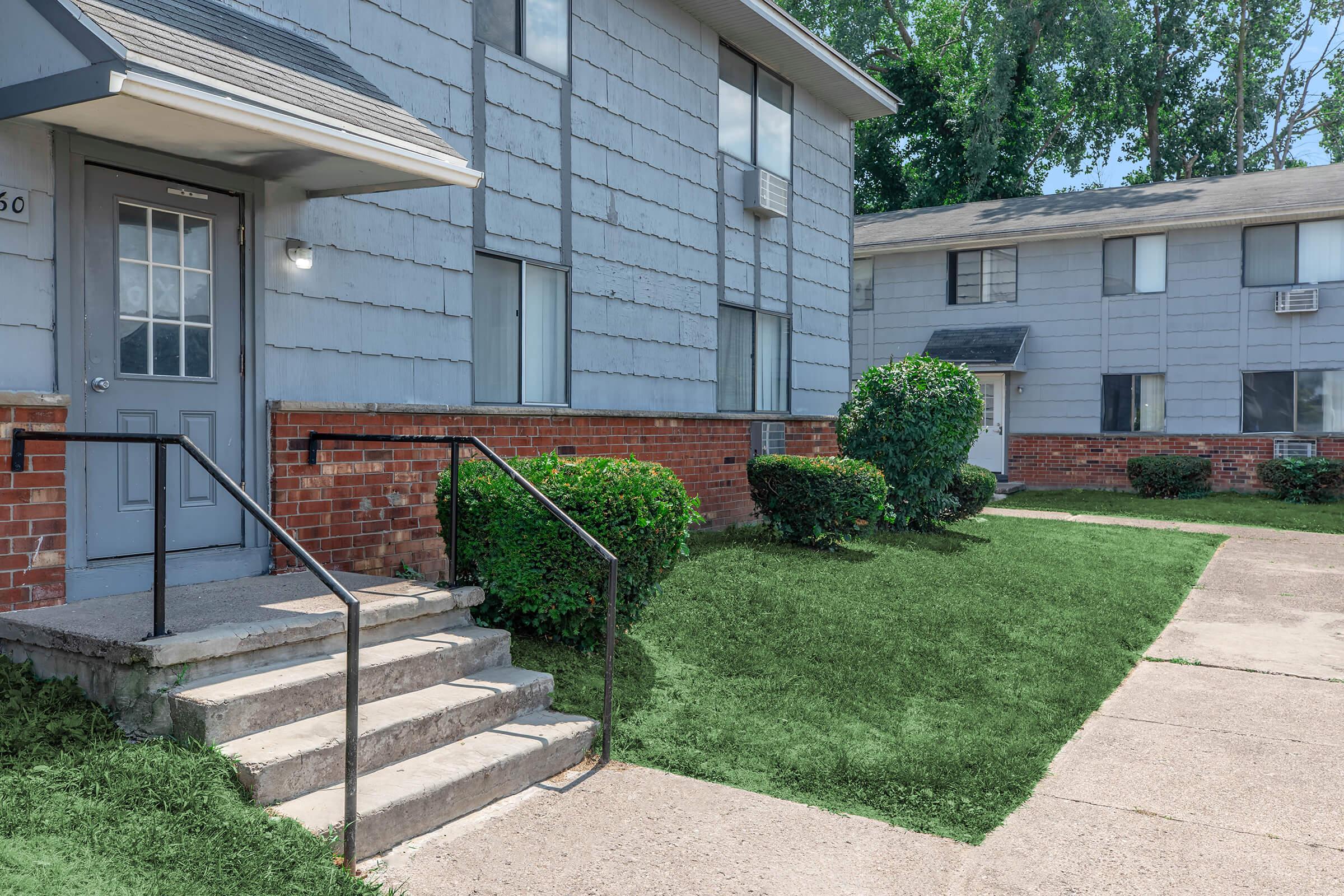 A view of an apartment complex featuring two buildings with gray siding and red brick accents. The foreground shows a set of concrete steps leading to a door, flanked by well-maintained bushes. A patch of green grass is visible on the ground. The scene is bright and sunny, suggesting a pleasant day.