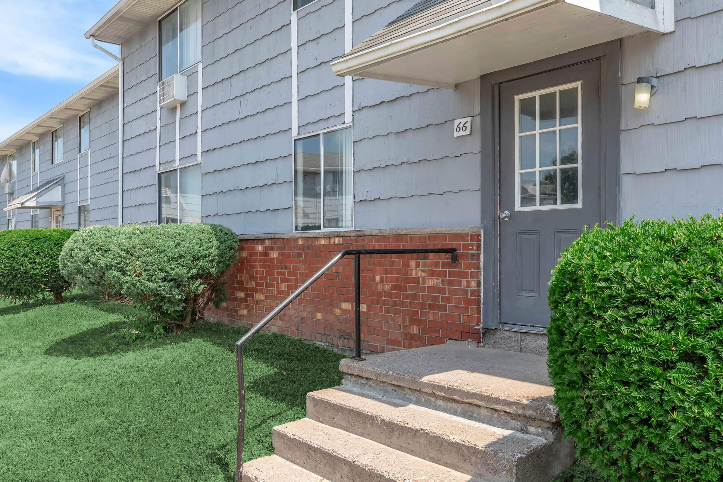 Exterior view of a multi-unit residential building featuring a gray exterior, a front door with glass panels, and concrete steps leading up to it. Lush, neatly trimmed shrubs flank the entrance, and a grassy area adds to the inviting atmosphere. The image captures a sunny day with clear skies.