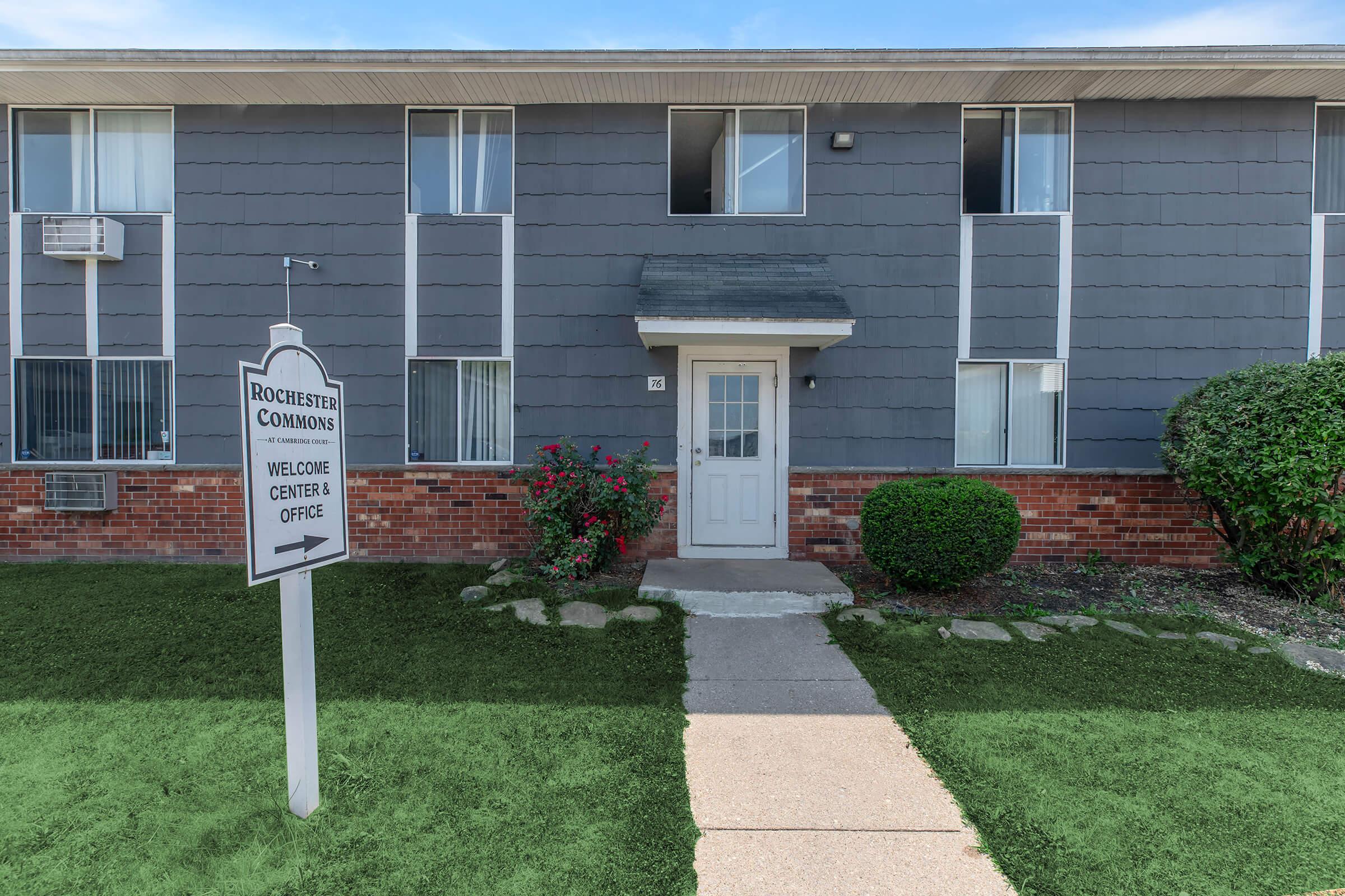Exterior view of a multi-unit residential building with a gray facade. In front, there's a sign directing to the welcome center office, and the walkway is bordered by manicured grass and small bushes. The building features several windows and a central entrance door.