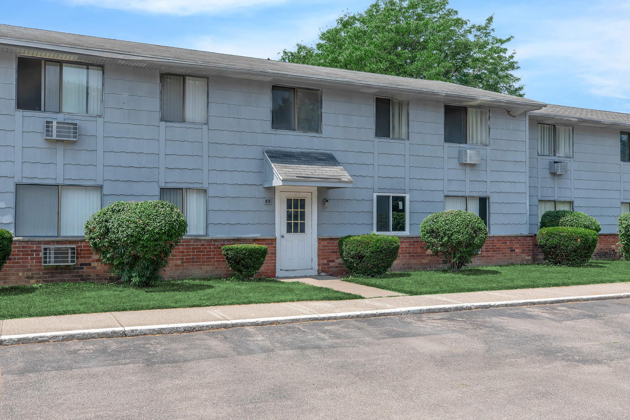 Two-story apartment building with a light gray exterior, featuring several windows and air conditioning units. The entrance has a white door and a small awning. Lush green bushes line the walkway, leading to the door, and a well-kept lawn is visible in front. A paved road runs along the building.