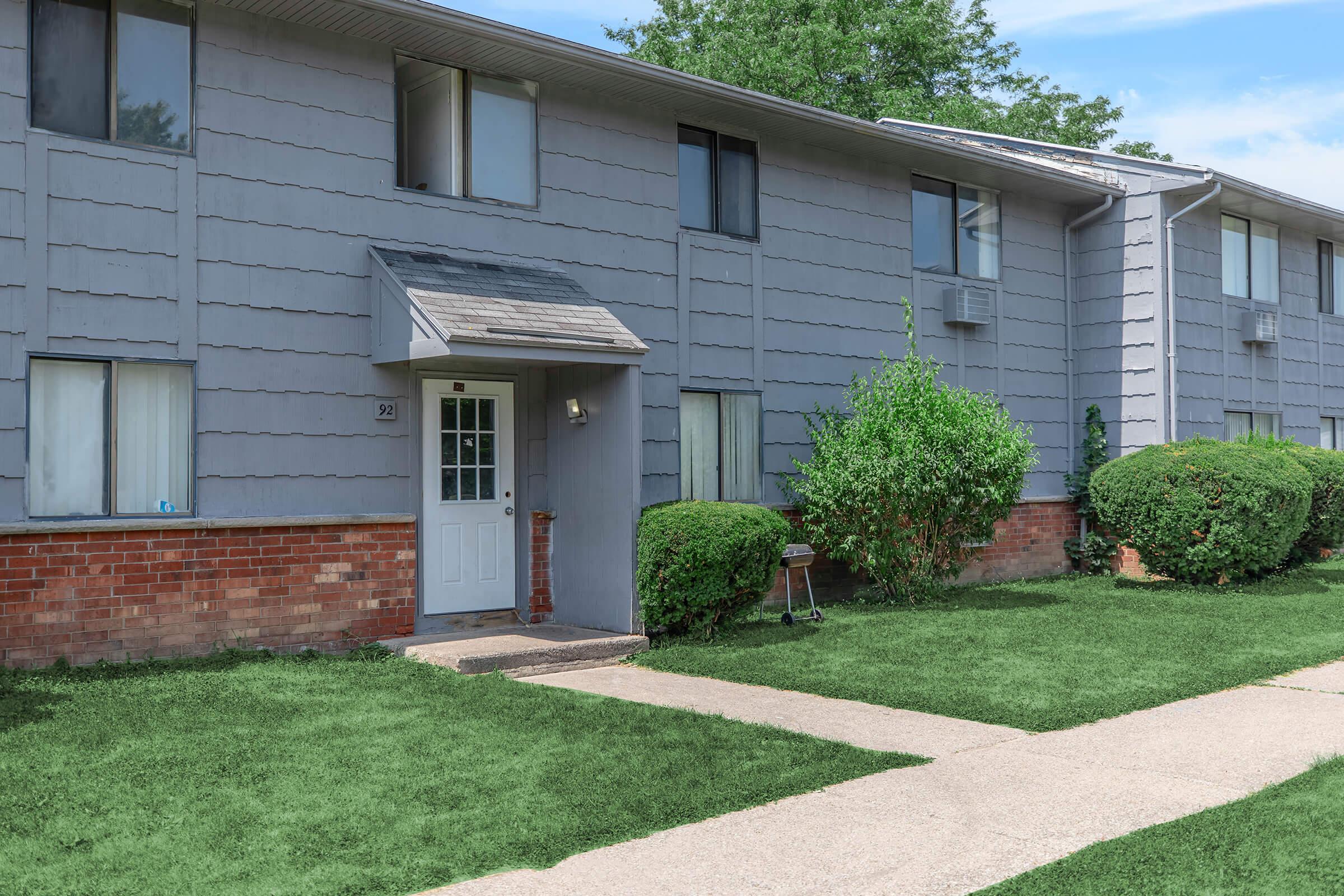 A residential building with a light gray exterior and brick accents. The entrance features a white door and a small overhang. Lush green grass surrounds the walkway leading to the door, with neatly trimmed bushes on either side. The building has several windows, some with air conditioning units. Trees and a blue sky are visible in the background.
